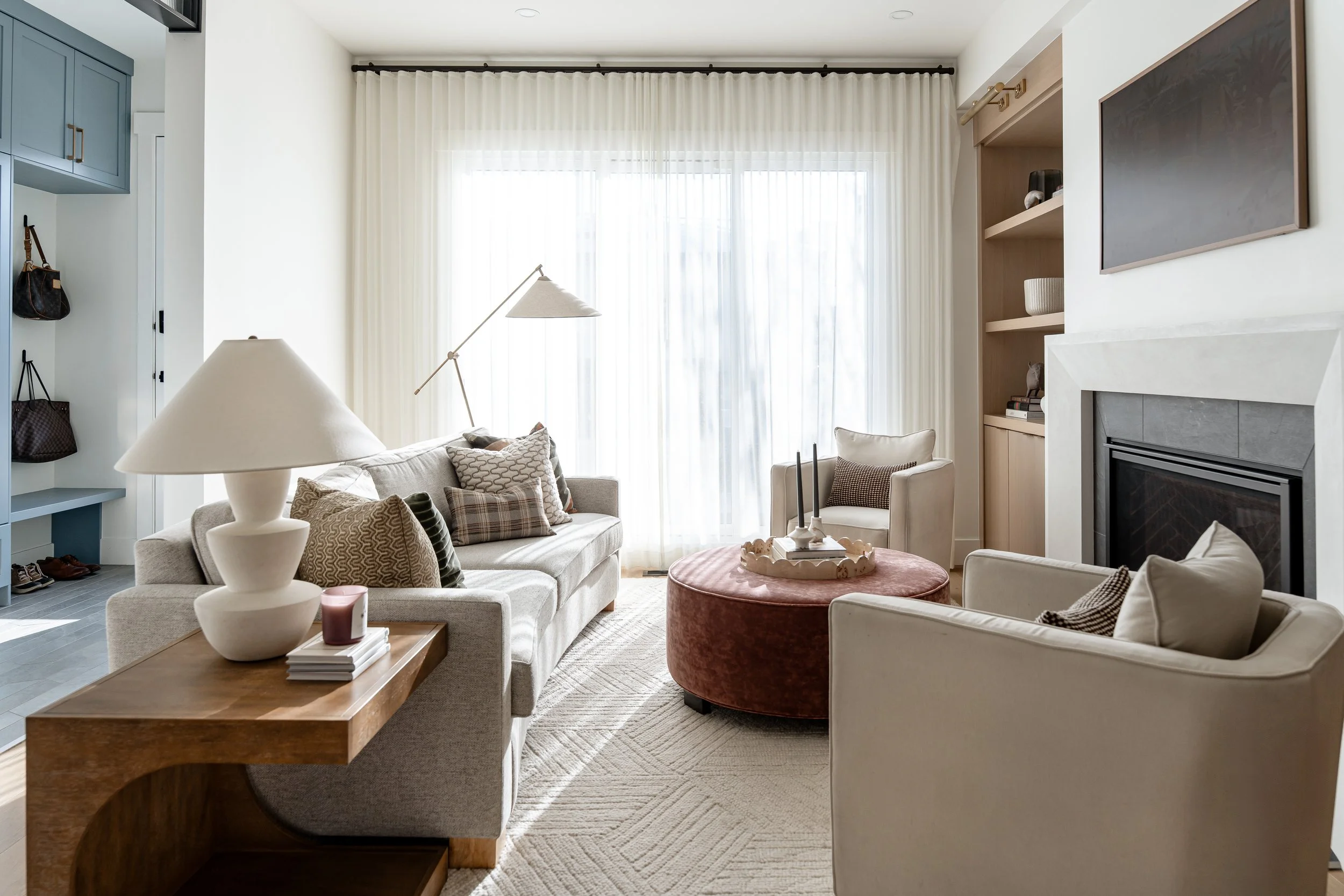 Living room with beige sofas, a wooden coffee table, a round ottoman, floor-to-ceiling curtains, and built-in shelves with decorative items.