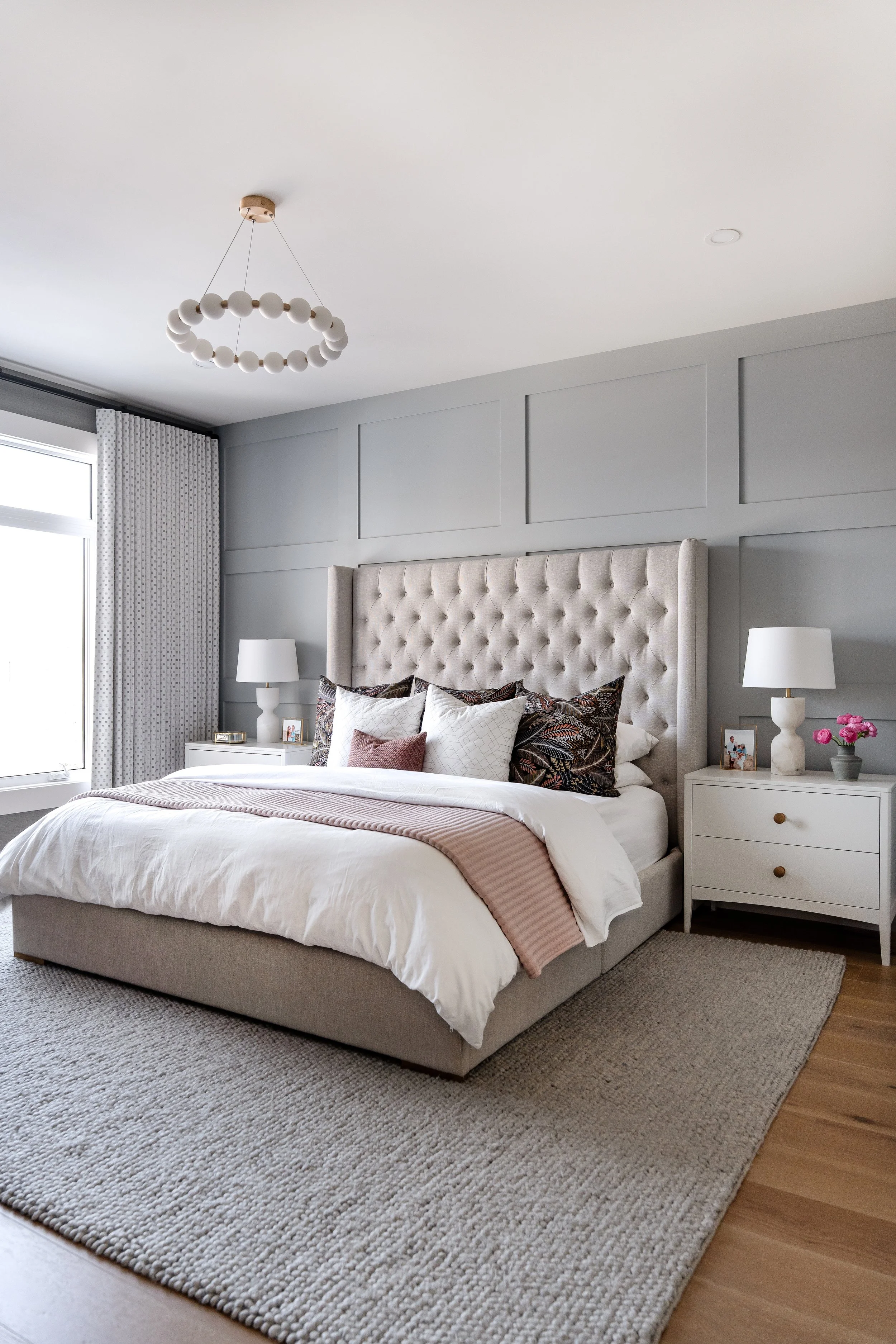 A bedroom with a large bed having a tufted headboard, white bedding, and decorative pillows, flanked by white nightstands with lamps, next to a window with curtains, a beige area rug, and a white ceiling with a modern chandelier.