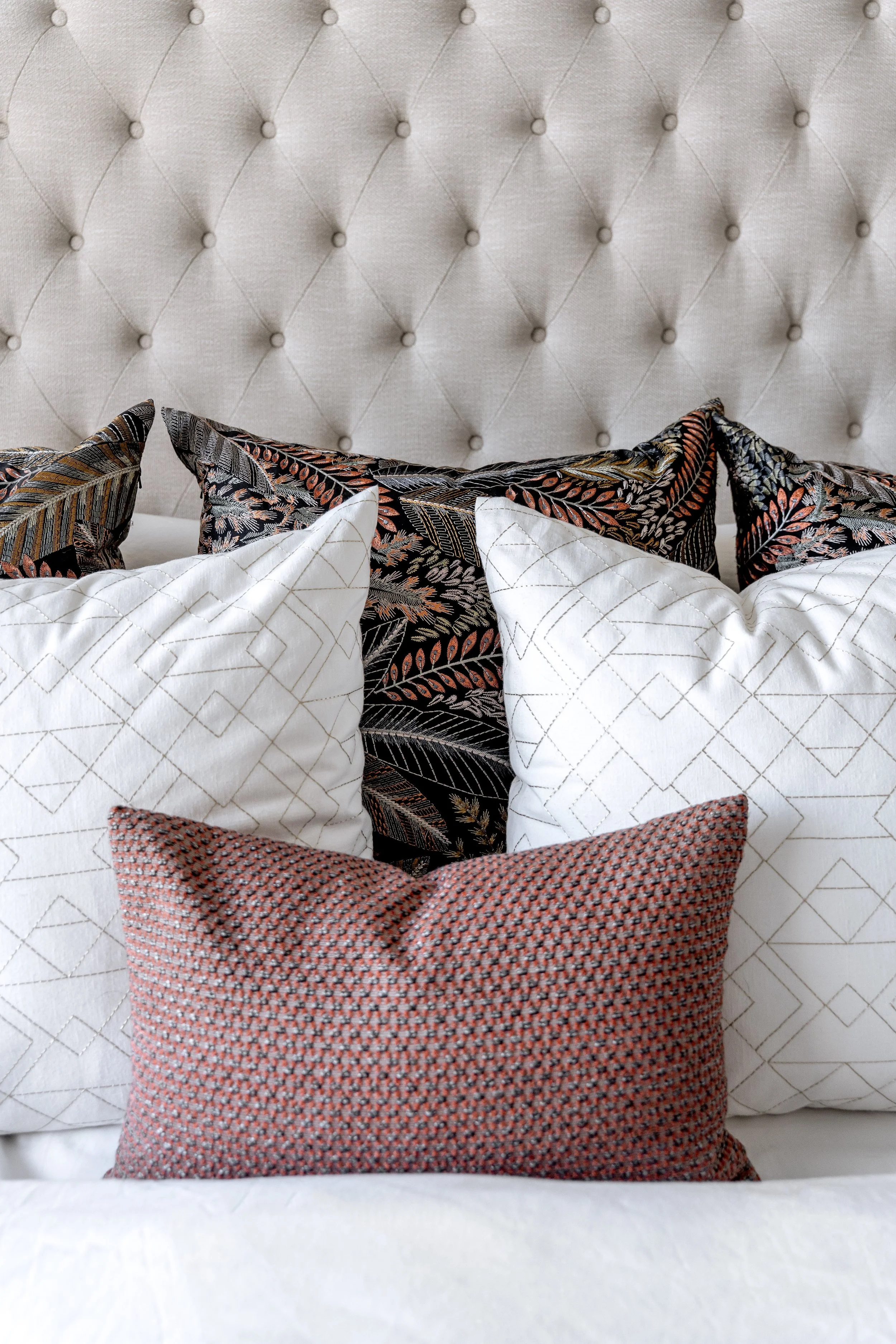 A bed with a white quilted cover and multiple decorative pillows, including two white pillows with gold lines, two black pillows with a floral pattern, and a red textured pillow. The bed features a tufted beige headboard with diamond stitching.