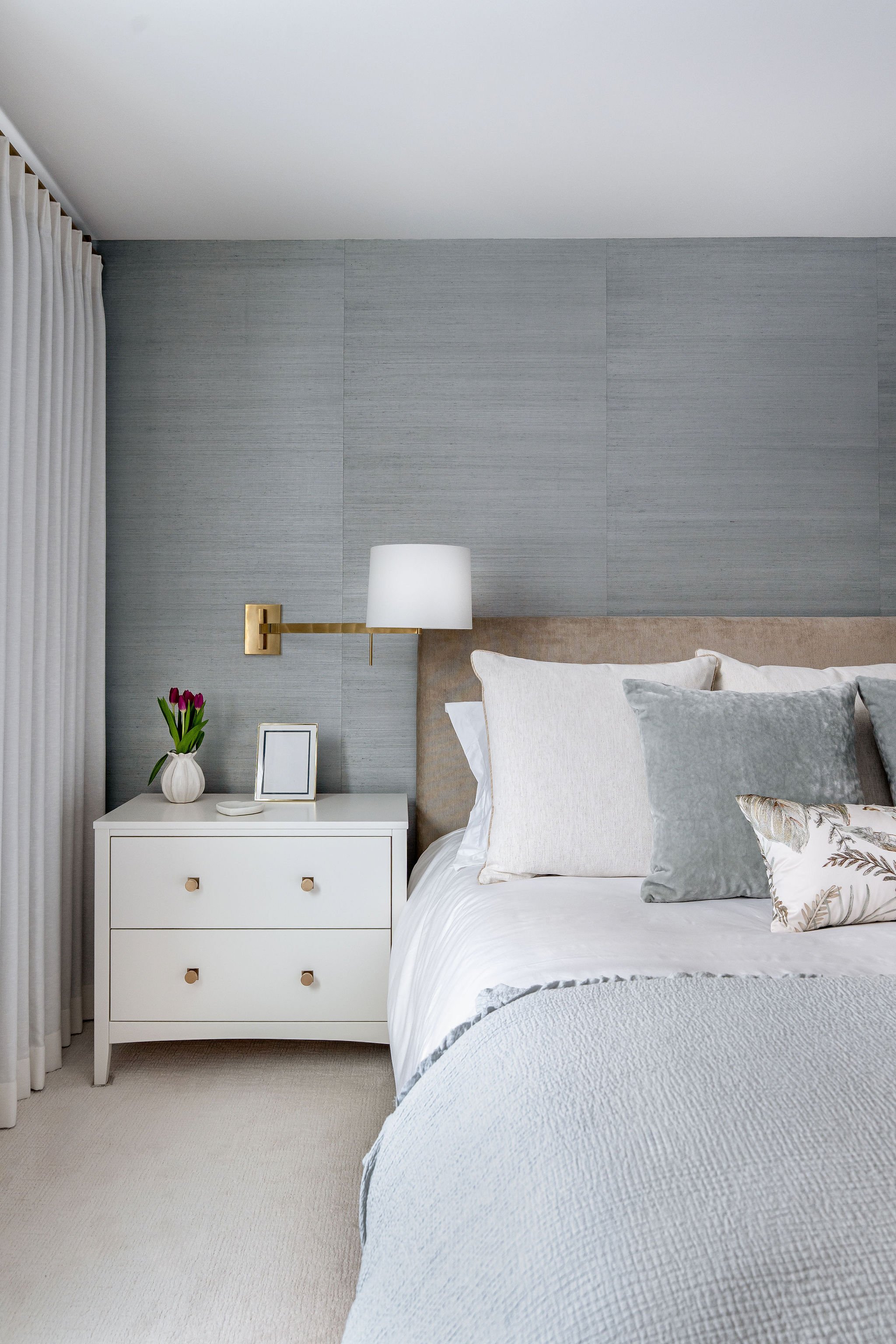 A bedroom with a white nightstand holding a vase of purple tulips, a picture frame, and a small white object. The bed has white bedding with multiple pillows, including a textured gray pillow and a decorative pillow with a leaf pattern. A wall-mounte