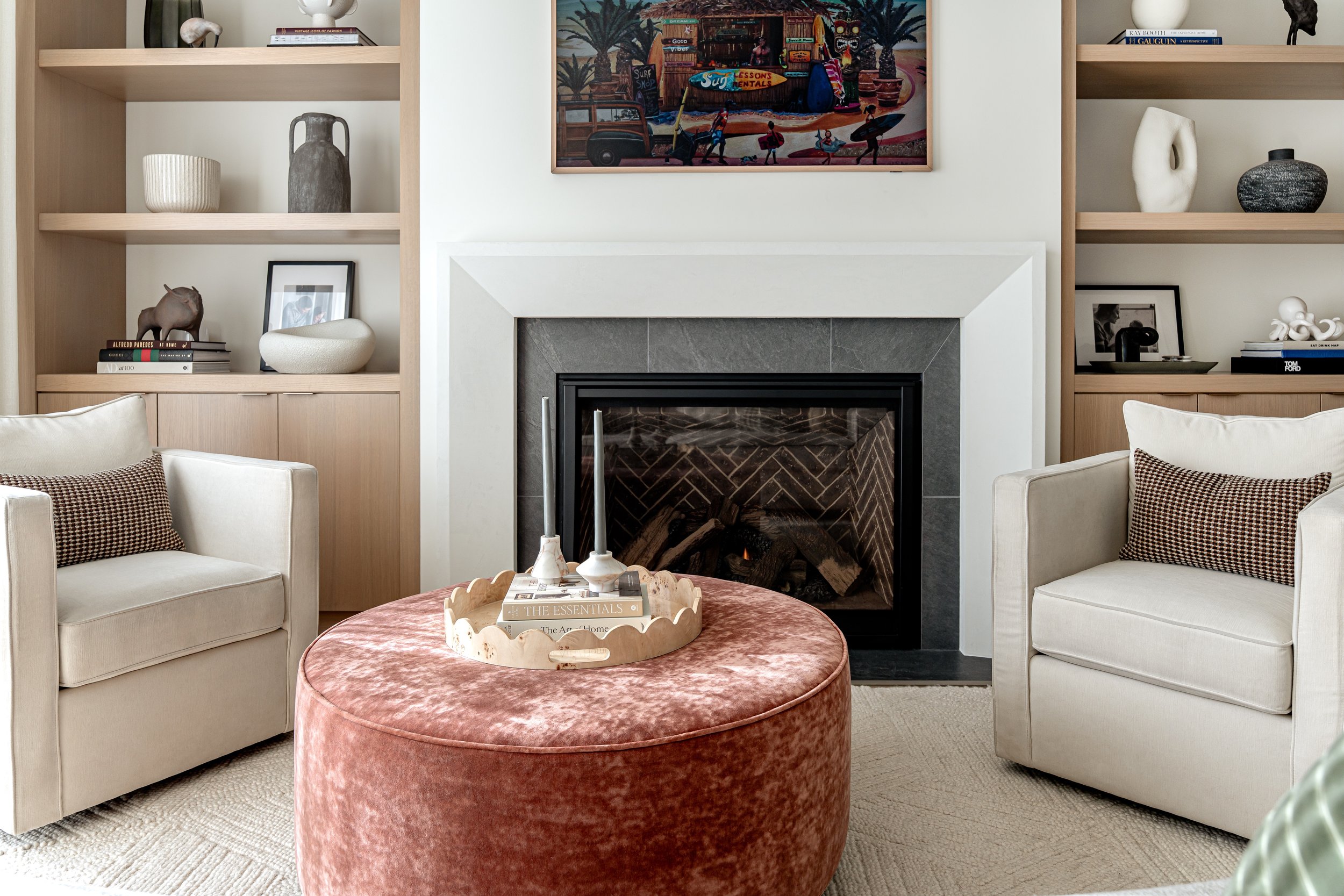 Living room with two cream-colored armchairs with patterned pillows on each side of a fireplace. A round, pink velvet ottoman with a decorative tray holding books and candles is in the center. On the shelves above the fireplace, there are decorative 