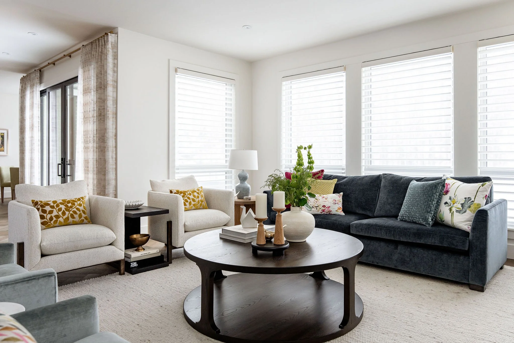 Living room with white walls, large windows with blinds, a black sofa with decorative pillows, beige armchairs with patterned pillows, a round wooden coffee table with decorative items, and a side table with a lamp and plants.
