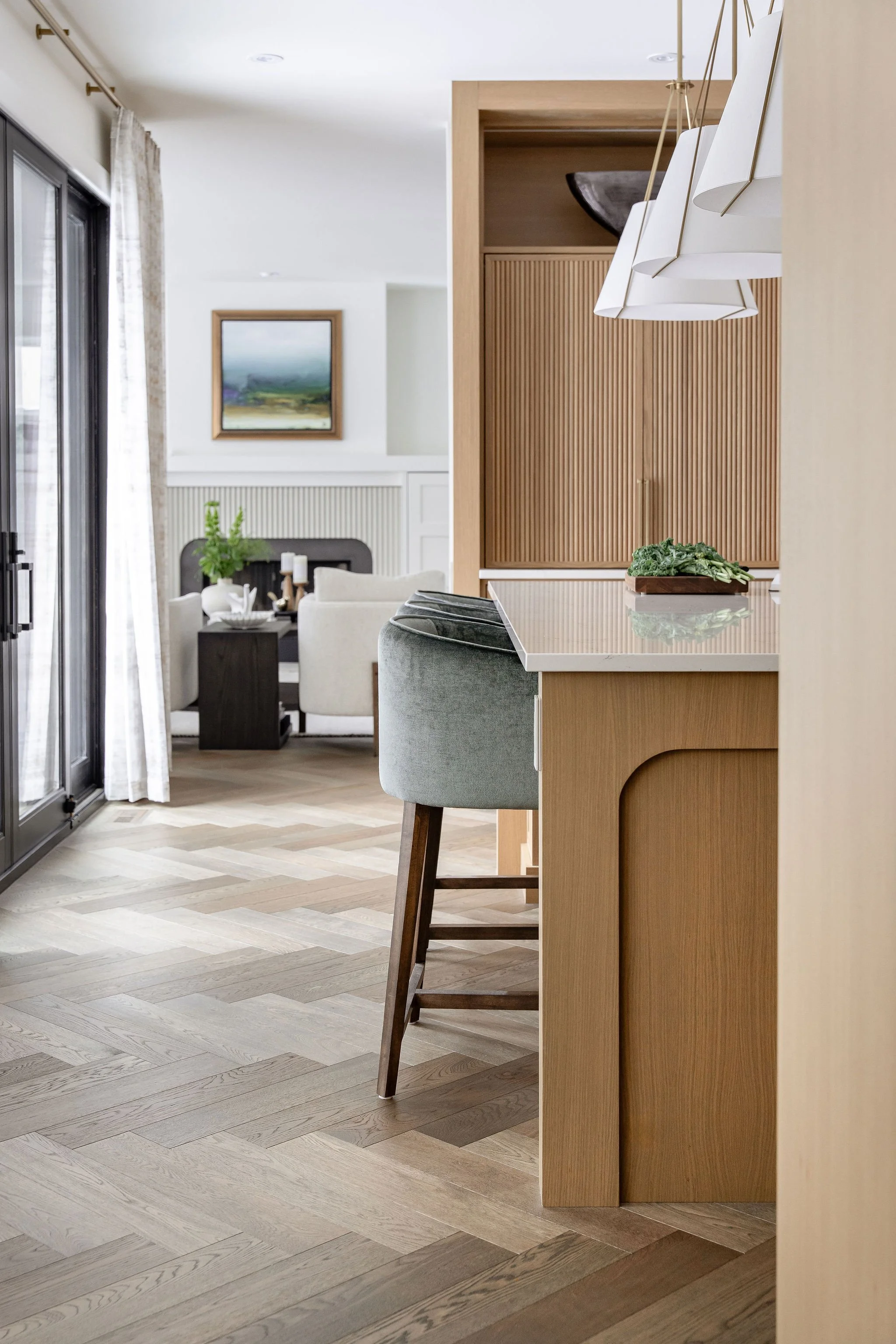 A modern kitchen with a light wood island counter, gray upholstered bar stool, and white hanging pendant lights. In the background, there is a dining area with white chairs, a dark table, and a wall with a framed landscape painting. The room has larg