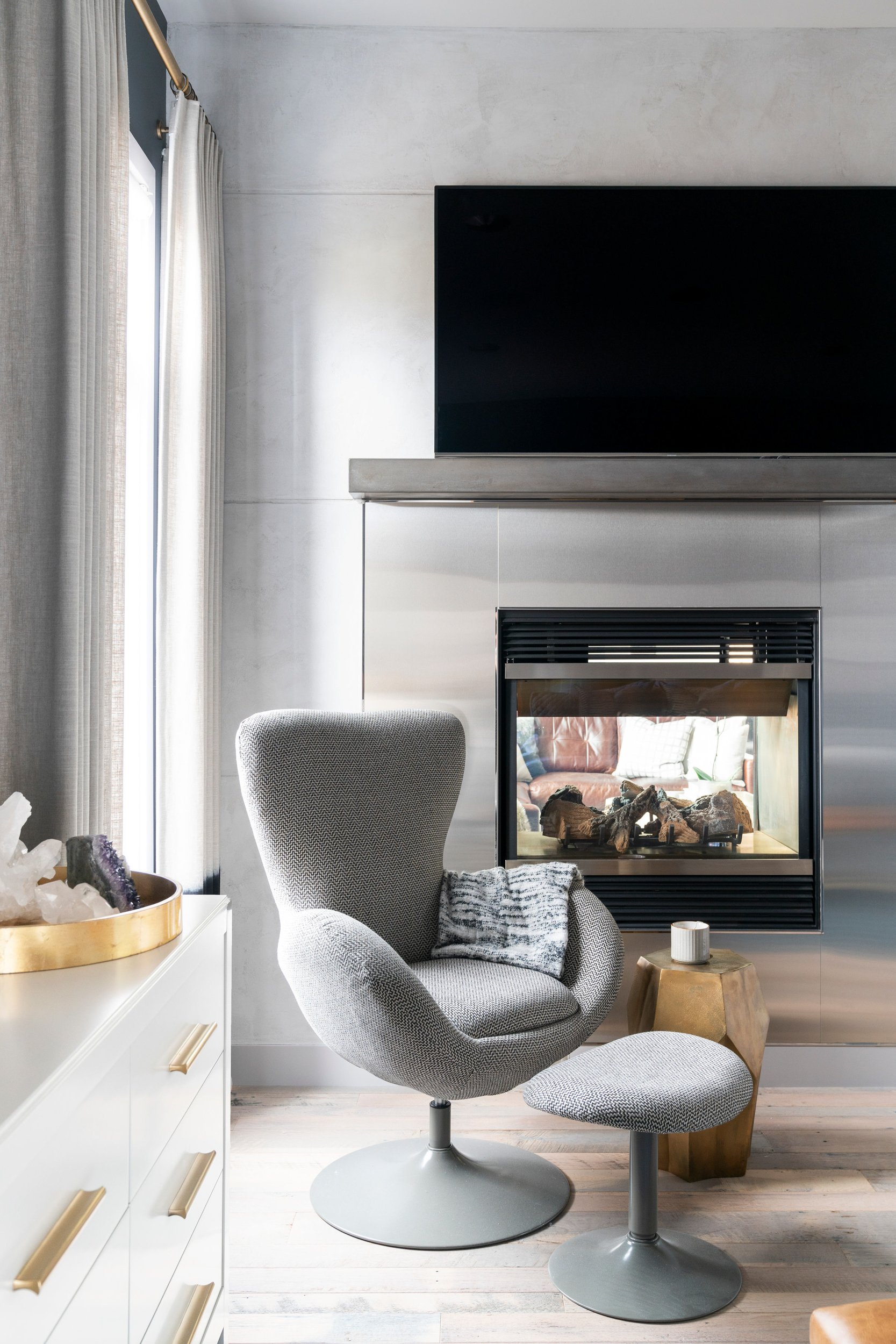 Modern living room with a gray swivel chair and matching ottoman in front of a fireplace. A TV is mounted above the fireplace. A white dresser with gold handles is on the left, featuring a decorative tray with crystals. Light-colored curtains cover t