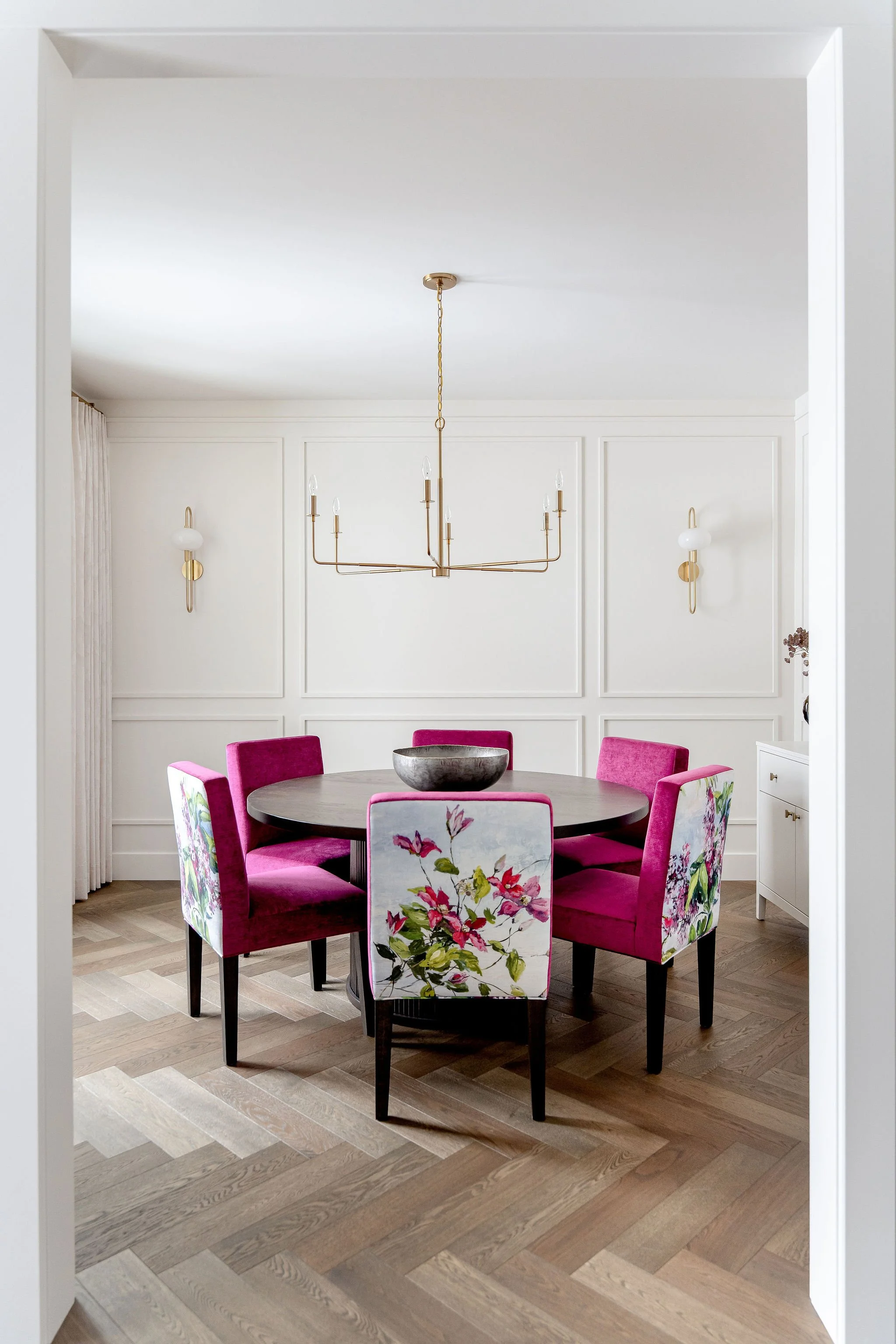 A dining room with a round dark wood table surrounded by six upholstered chairs, five of which are deep pink velvet, and one with floral upholstery featuring pink flowers and green leaves. On the table is a large gray bowl. The walls are white with d