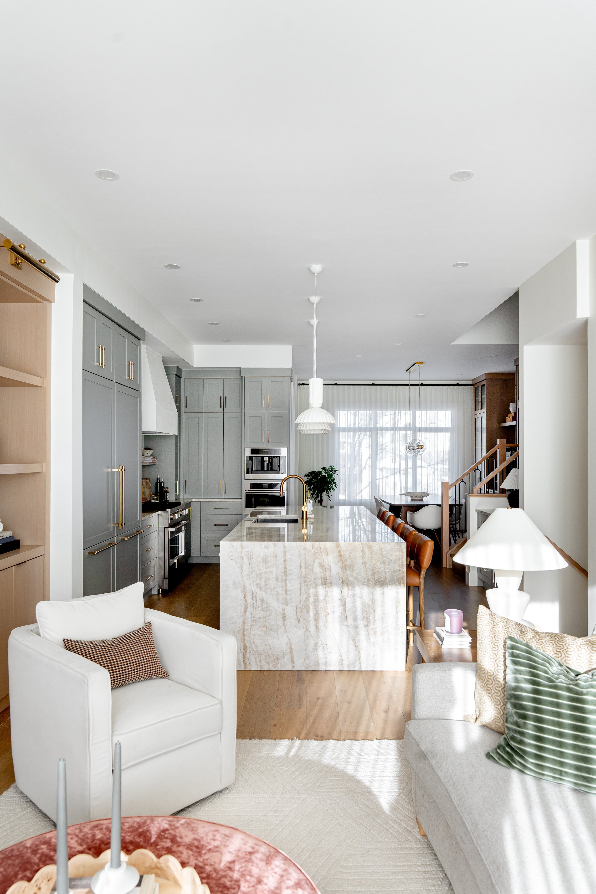 A modern, bright living space and kitchen with light-colored walls, built-in cabinets, a marble island, and a sitting area featuring a white armchair and a secondary gray sofa with varied cushions. Large windows with sheer curtains allow natural ligh