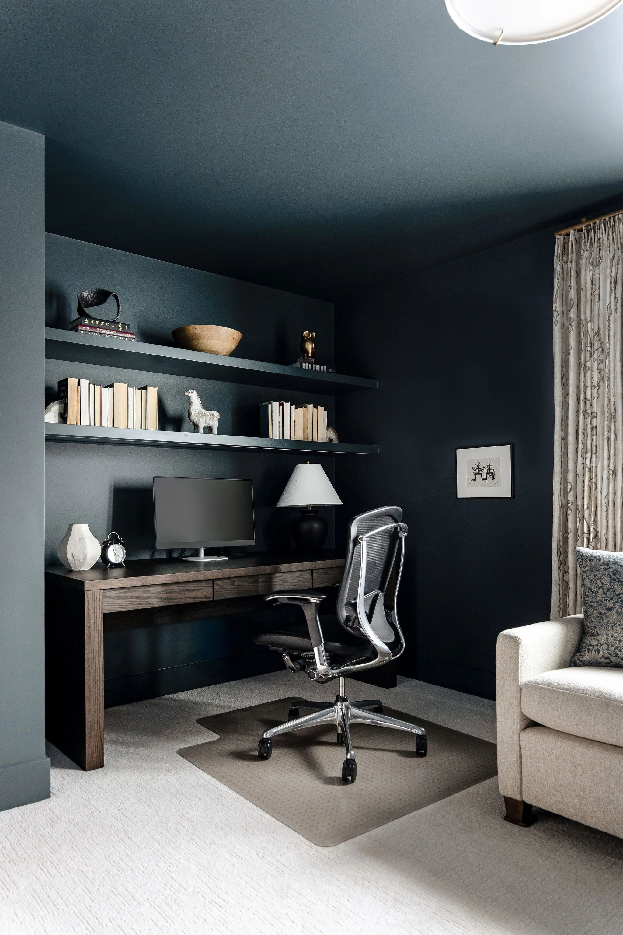 A home office with a wooden desk, black and chrome ergonomic office chair, computer monitor, and desk lamp. Shelves above the desk hold books, a wooden bowl, and decorative items. To the right, a beige armchair with a patterned pillow is next to a wi