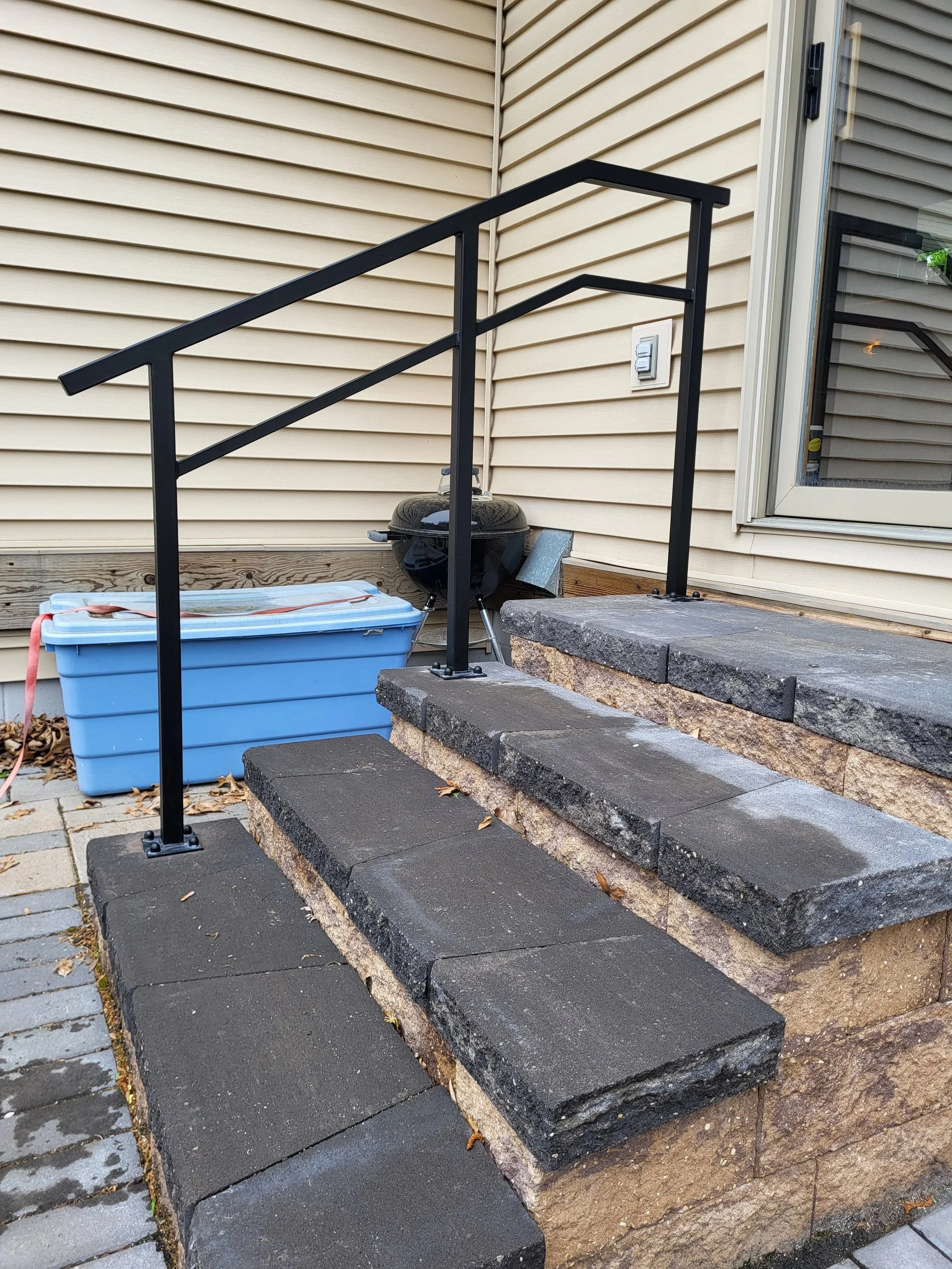 Outside residential stairs with black metal railing, leading up to a patio area with a sliding door, and a charcoal grill nearby; a blue plastic storage container and potted plants in the background.