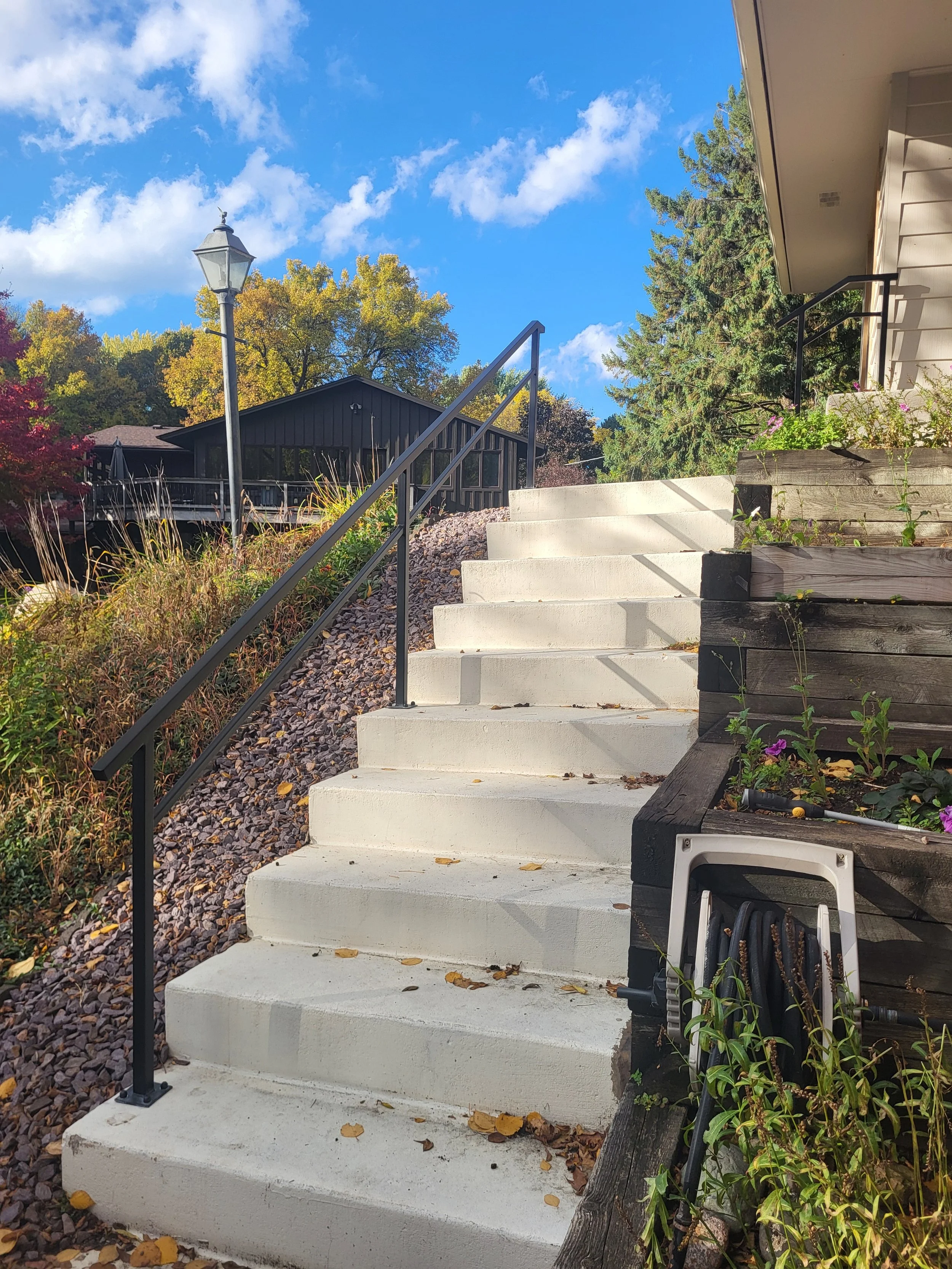 Exterior view of concrete stairs with black metal railing next to landscaped garden with plants and flowers, leading up to a house under a blue sky with scattered clouds.