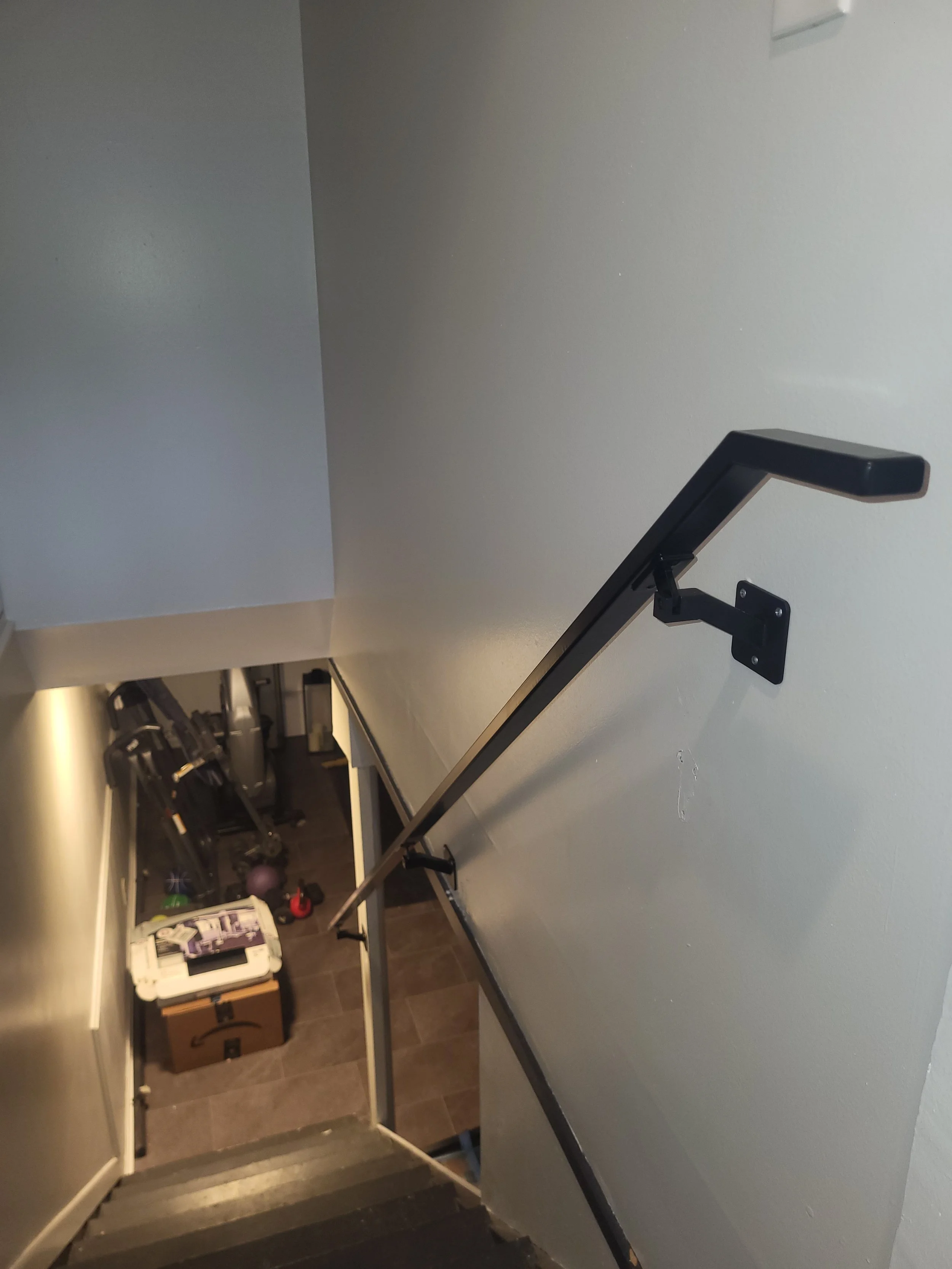 View of a staircase from the top, showing the handrail and the stairs leading down to a small landing area with some storage boxes, exercise equipment, and a medicine ball.