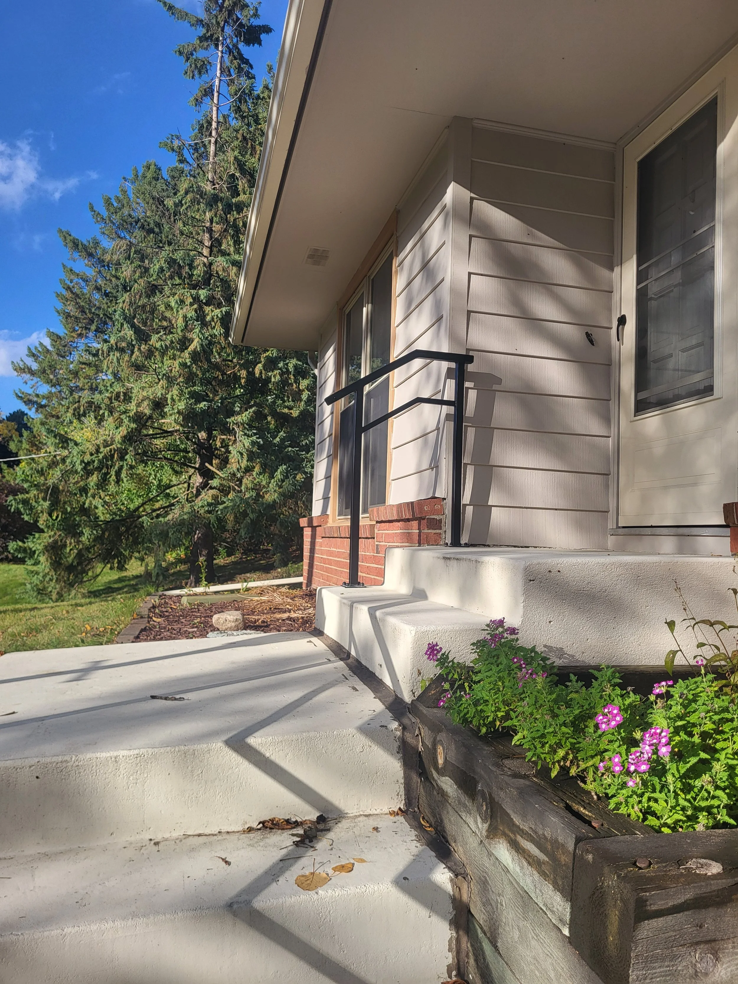 The image shows the front porch of a house with white steps, a black metal railing, and a flower bed with purple flowers. There are trees, a blue sky, and part of the house's exterior wall with windows and a door.