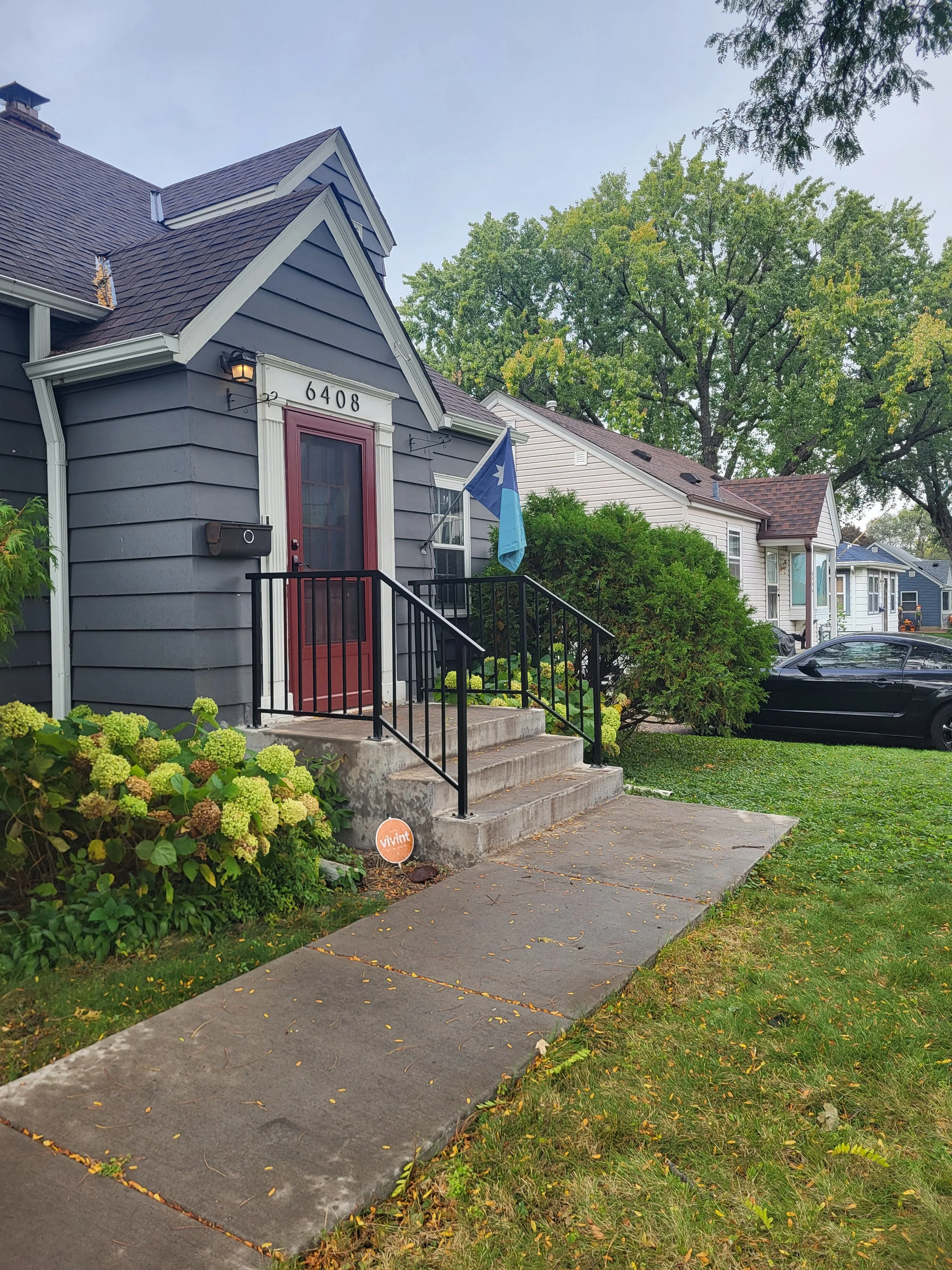 A house with gray siding, a red front door, a porch with black railing, and a Texas flag hanging outside. There is a concrete walkway leading to the stairs and a black car parked in the driveway. Green shrubs and trees are surrounding the house.