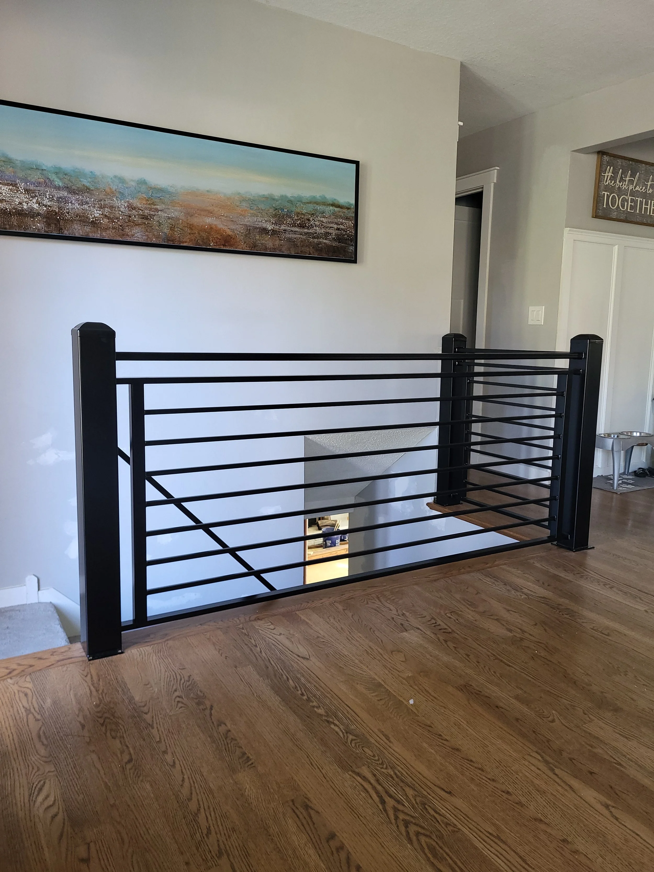 Interior view of a house showing a black metal railing overlooking a staircase and living area, with a large landscape painting on the wall and part of a hallway visible.