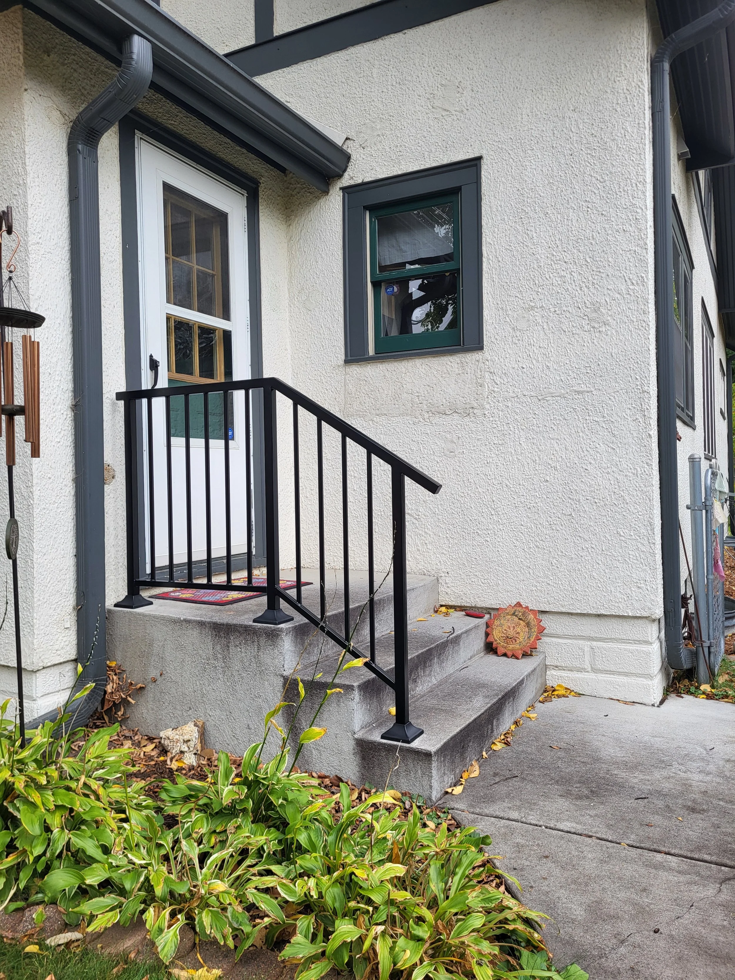 Front entrance of a house with a small concrete staircase, black metal railing, white door, and a small window above the stairs. There is a decorative item on the ground to the right of the stairs and plants in the garden bed at the bottom of the ima