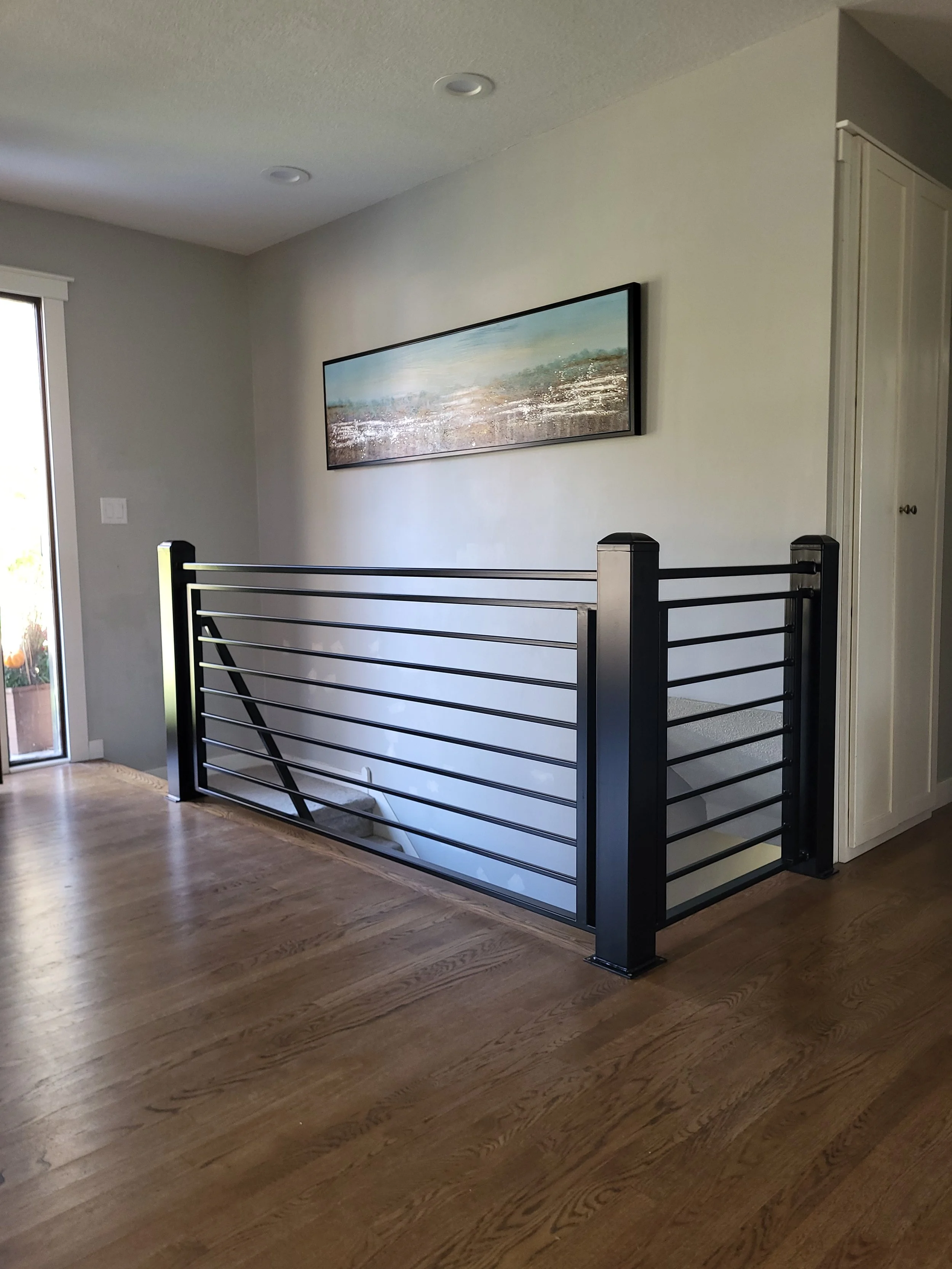 Interior of a home with a black metal staircase railing, wooden flooring, a sliding glass door to the left, a framed landscape painting on the wall, and a white cabinet on the right.