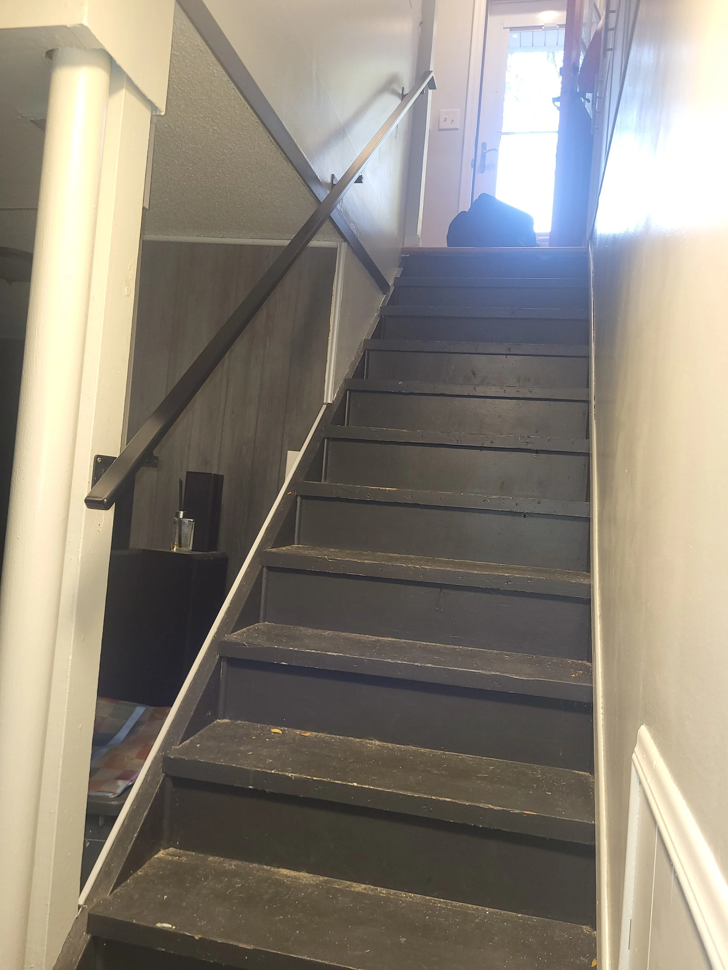 Interior view of a staircase with black steps and a black handrail, leading up to a door with glass window panels. Part of an indoor wall is visible, and sunlight is coming through the door.