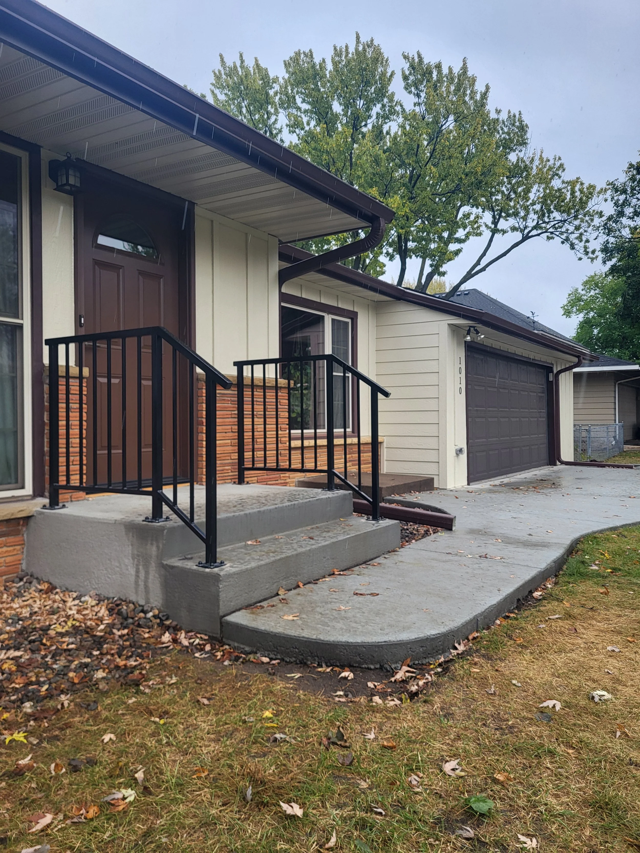 The front entrance of a house with a concrete porch, black metal railing, brown door, and beige siding, with a driveway leading to a garage. There are green trees and fallen leaves on the ground.