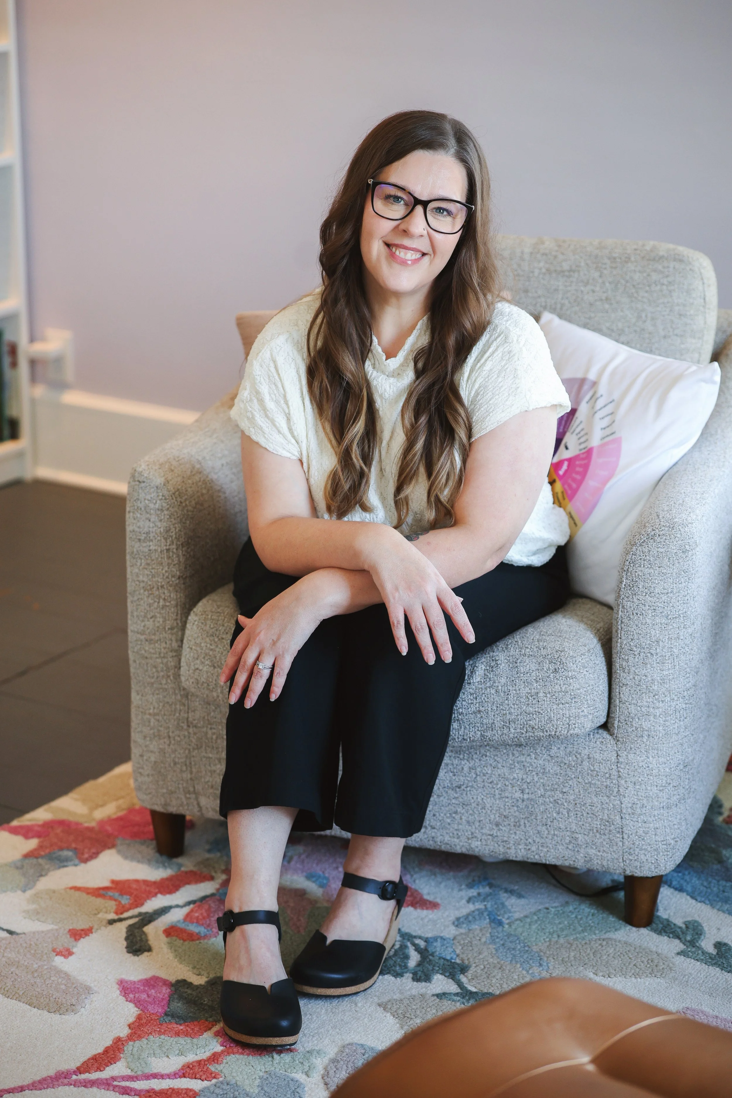 A woman and therapist Melissa Sichting with long hair and earrings smiling, wearing a striped sweater, sitting on a bench inside a room near a window.