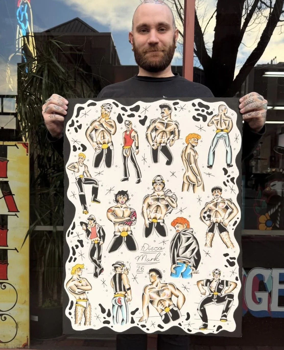A man with a beard and tattoos holding an illustrated poster featuring various cartoon-style male characters in different poses, all shirtless and styled in retro or vintage fashion.
