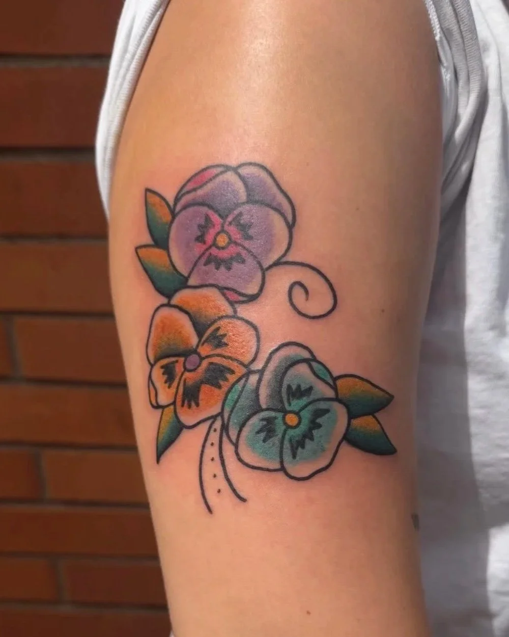 Colorful tattoo of three pansy flowers with green leaves on a person's upper arm.