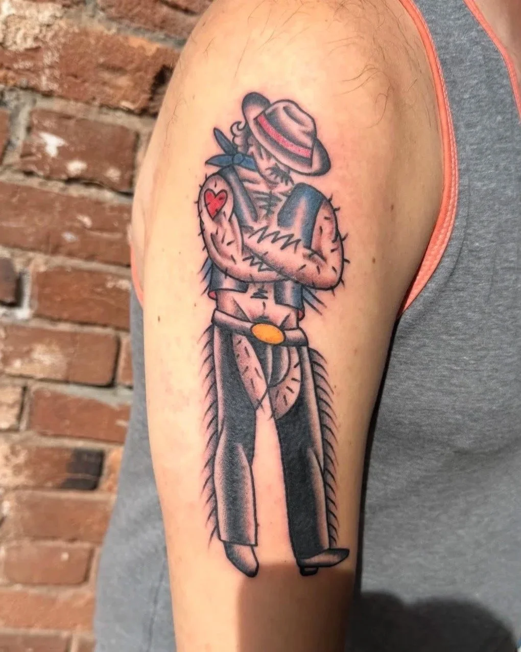 Tattoo of a cactus person dressed as a cowboy, wearing a hat, with a heart-shaped tattoo on the arm, and standing against a brick wall.
