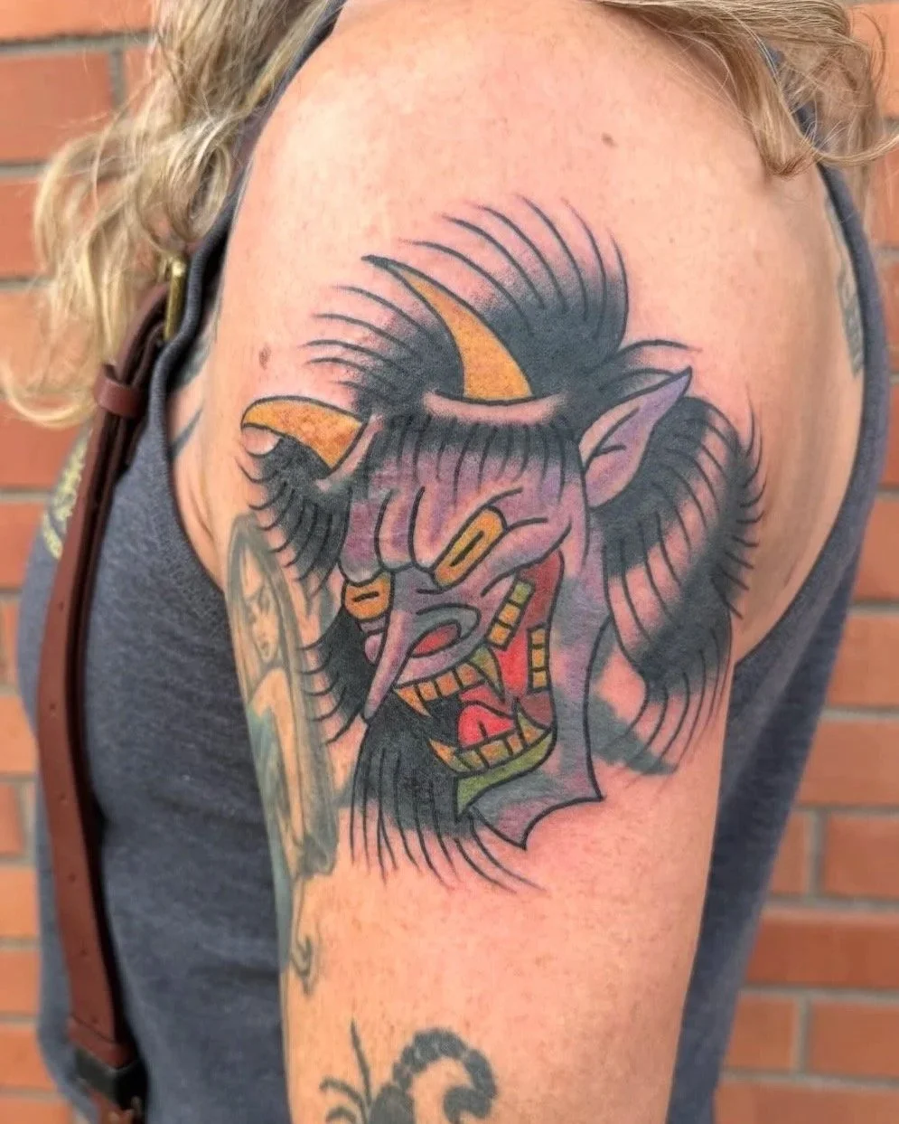 Tattoo of a menacing demon or gargoyle with horns and long hair on a person's upper arm, with a brick wall in the background.
