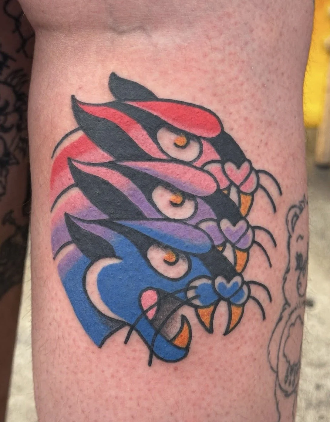 Tattoo of three colorful cats with hearts on their chests, arranged vertically, with a rainbow gradient in the background.