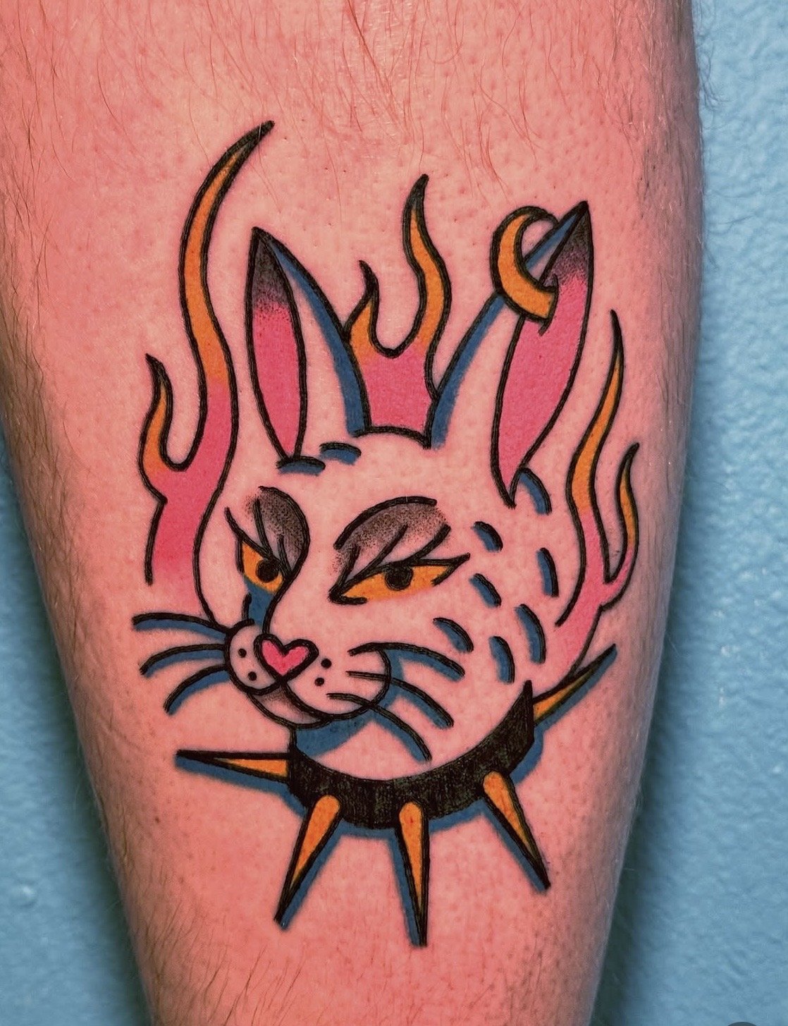 Tattoo of a stylized cat with flames around it, featuring a spiked collar, and a woman with long hair.