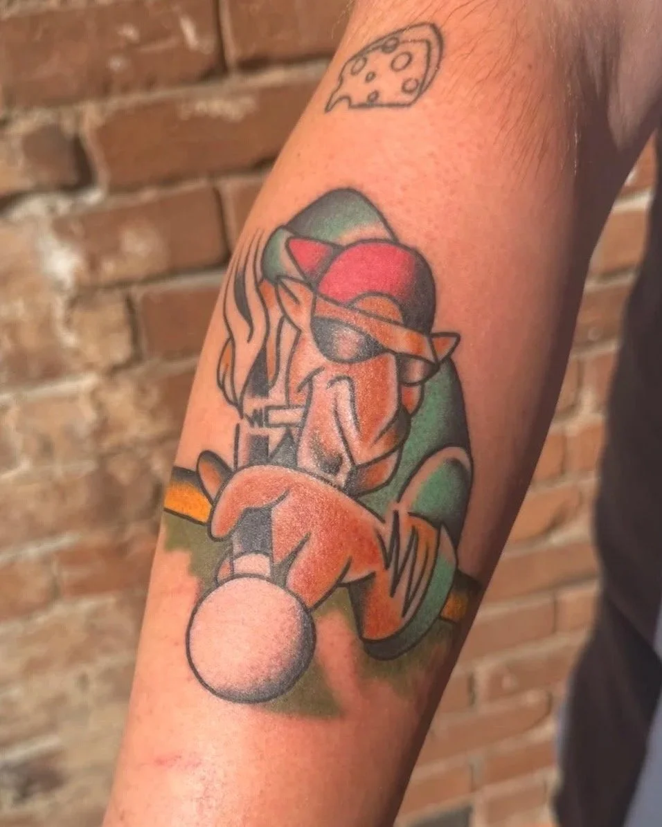 A colorful tattoo on a person's forearm features a cartoon-style dog wearing sunglasses and a red cap, sitting next to a large tennis ball. Above the dog, there is a line-drawing of a slice of cheese. The background includes a green and orange color scheme.