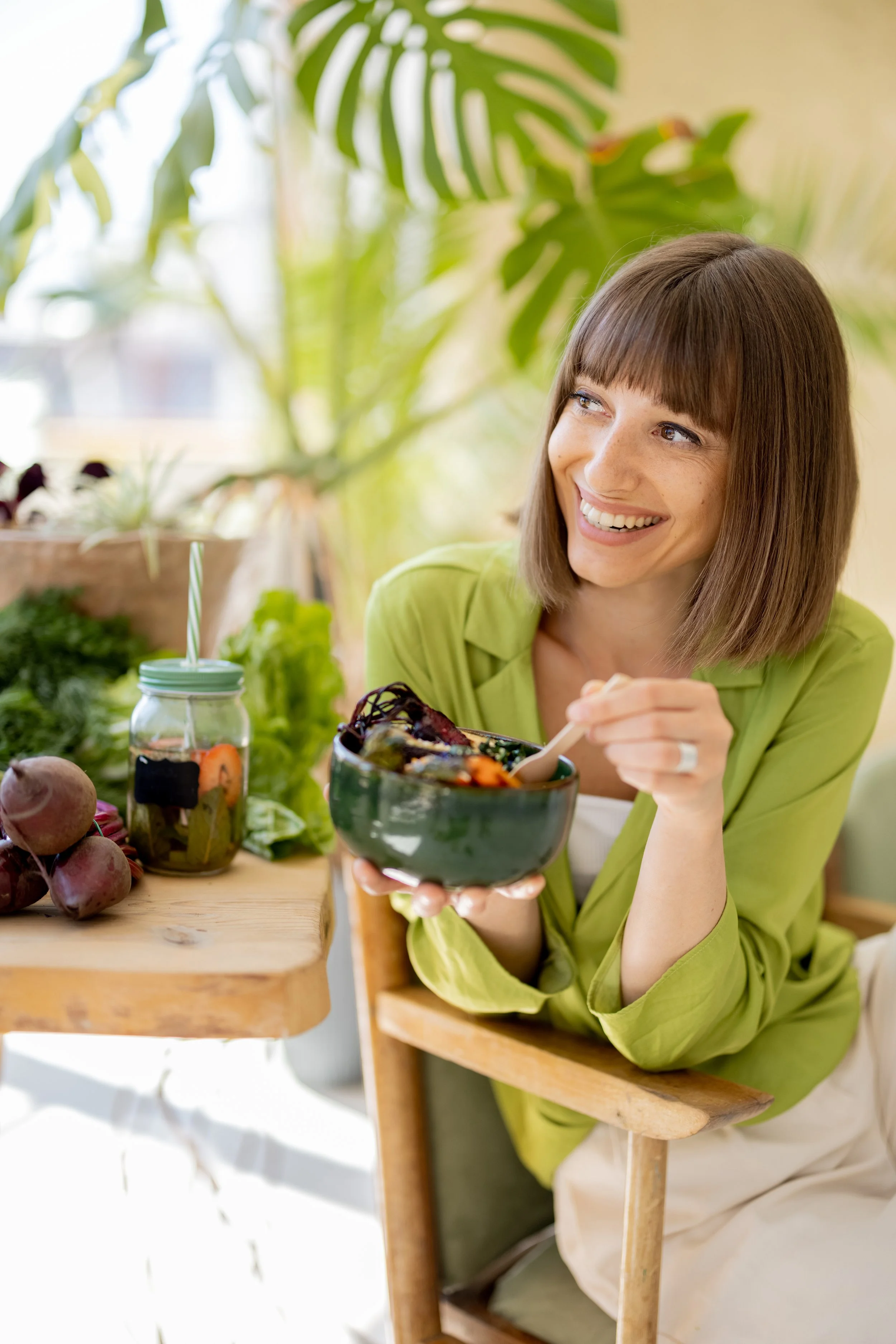 A woman with brown hair and freckles smiles while holding a black bowl of salad at a wooden table. There are various vegetables and a jar with a straw on the table, with a large green plant in the background.