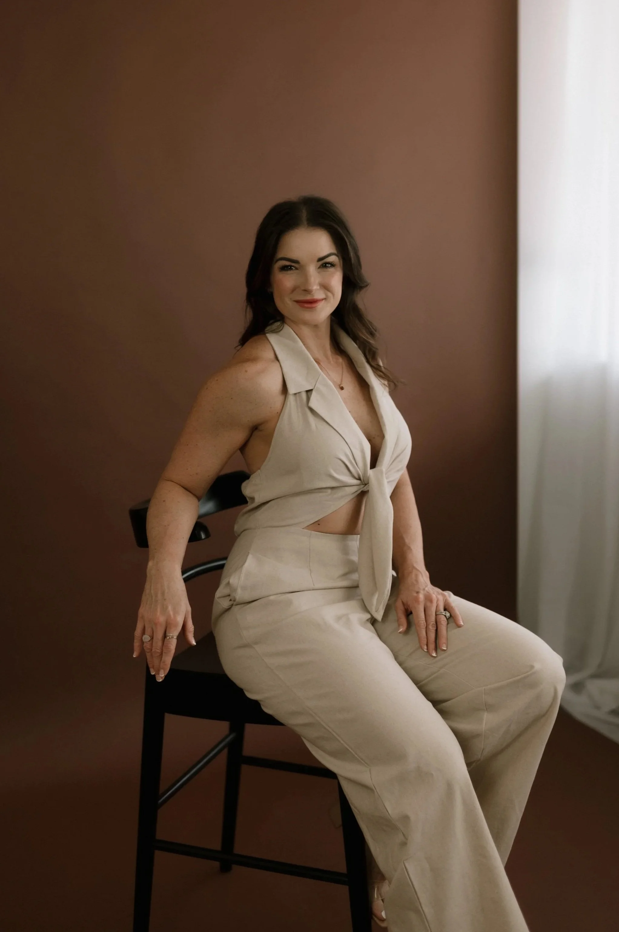 A woman with brown hair and a beige sleeveless top tied at the waist, sitting on a black chair against a brown wall, smiling at the camera.