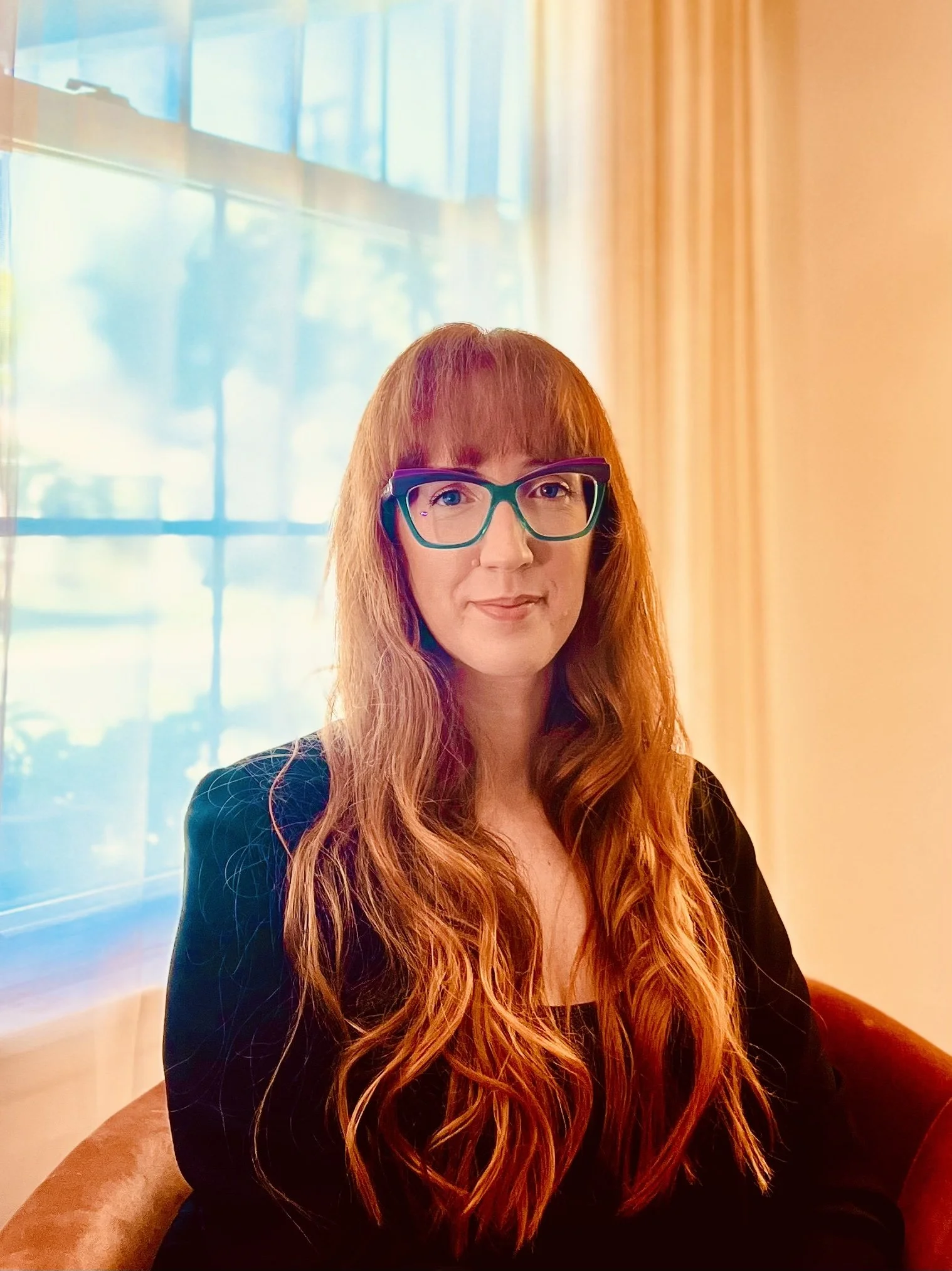 A woman with long wavy red hair and teal-rimmed glasses, sitting in front of a window with sheer curtains, wearing a black top.