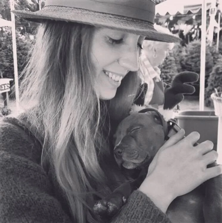 A woman in a wide-brimmed hat and sweater is smiling and hugging a sleeping puppy. The photo is in black and white and appears to be taken outdoors, possibly at a fair or outdoor market.