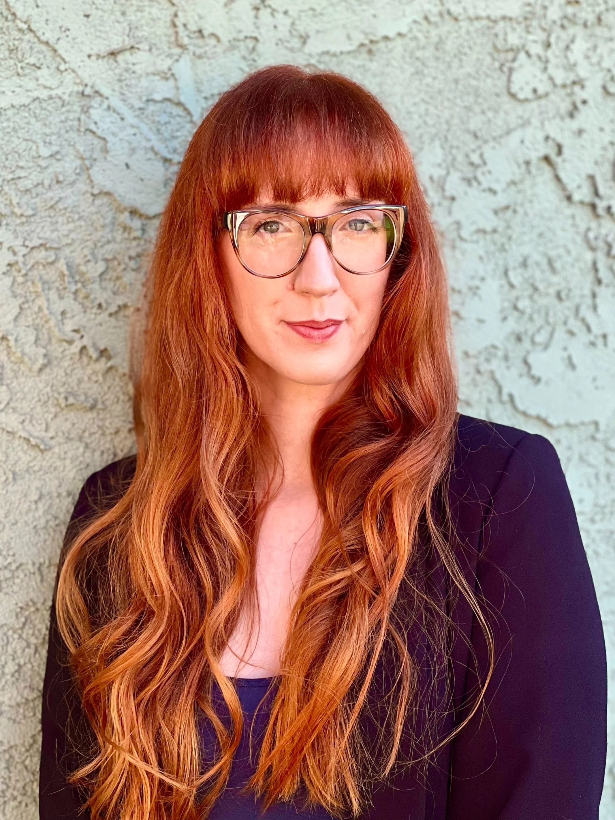 A woman with long wavy red hair, wearing glasses and a black blazer, standing against a textured light-colored wall.