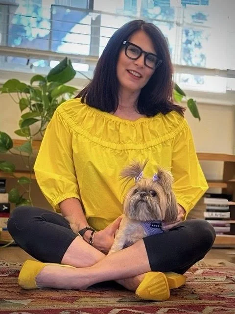 A woman with dark hair, glasses, and a yellow blouse sitting cross-legged on a rug with a small dog on her lap, in a room with plants and books.