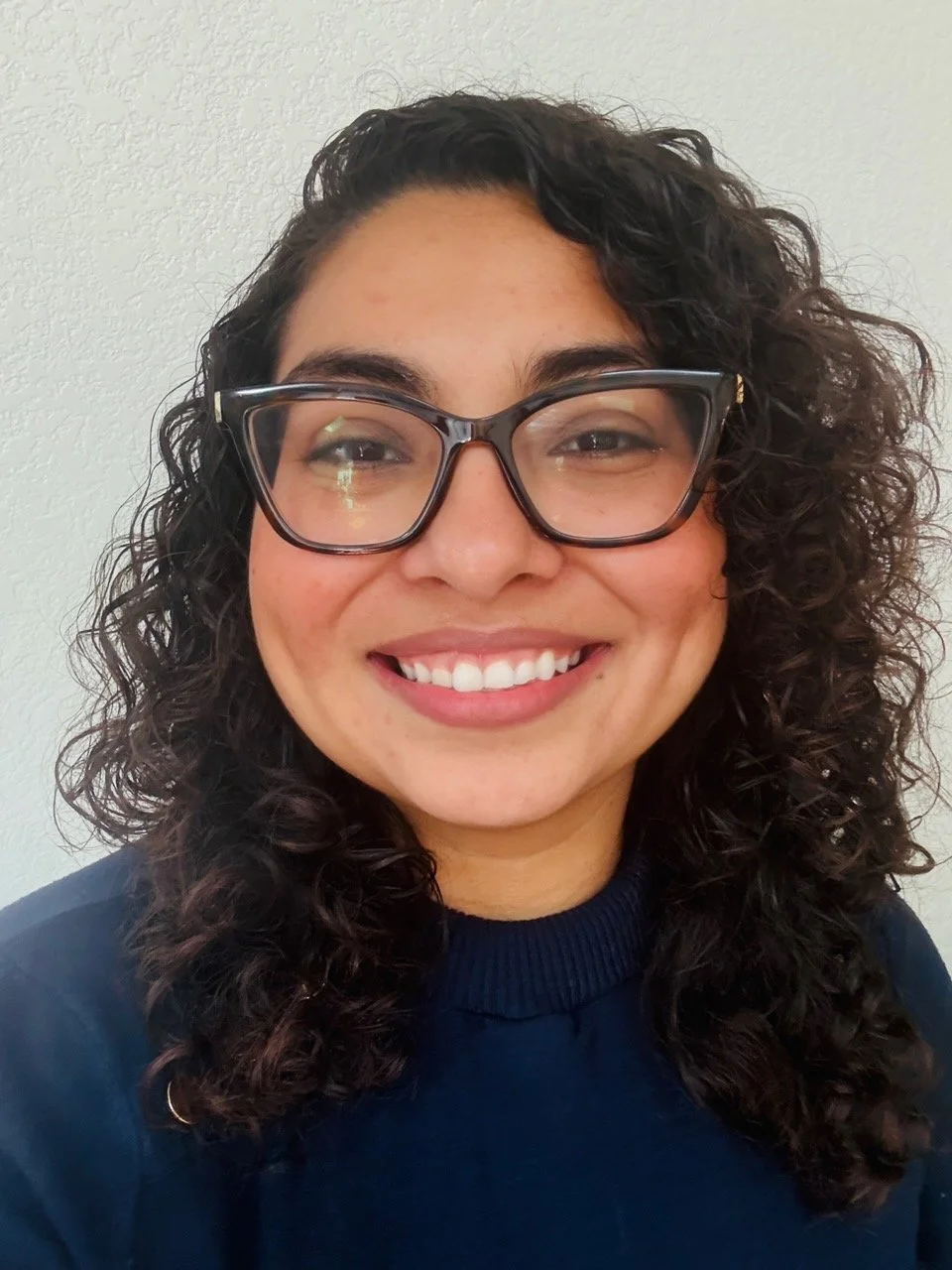 A woman with curly dark hair, glasses, and a bright smile wearing a dark sweater, standing against a plain light-colored wall.