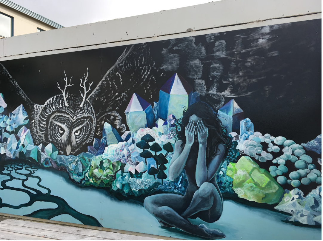 A mural depicting a woman with long dark hair, sitting with knees drawn up, covering her face with her hands. Behind her are various large, colorful, and iridescent crystals and gemstones, with a black and white owl with antlers in the background, and a dark sky with clouds. A winding river flows in front of the woman and the crystalline landscape.