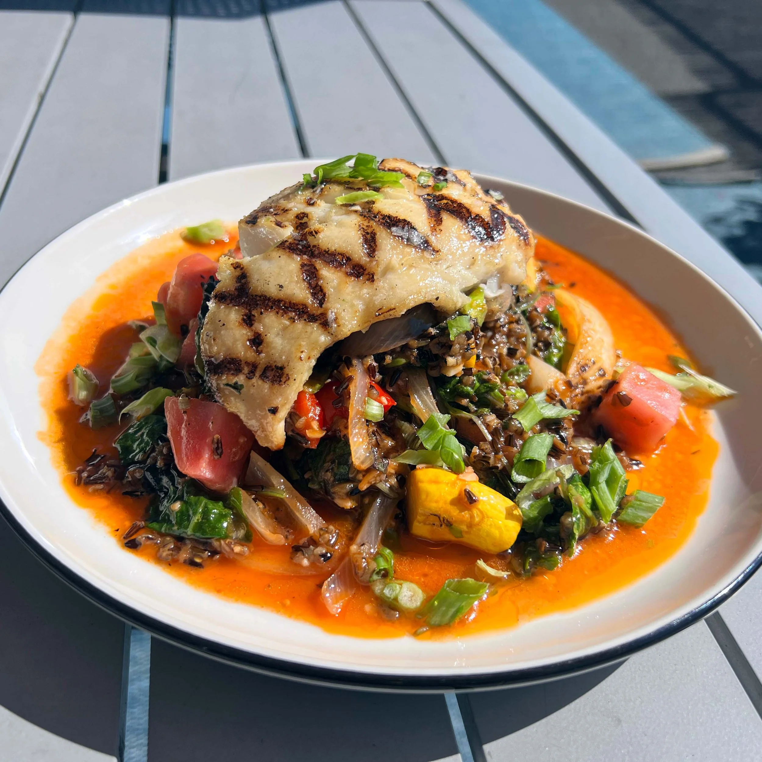 Grilled fish fillet on top of a vegetable medley in a spicy broth with chopped green onions