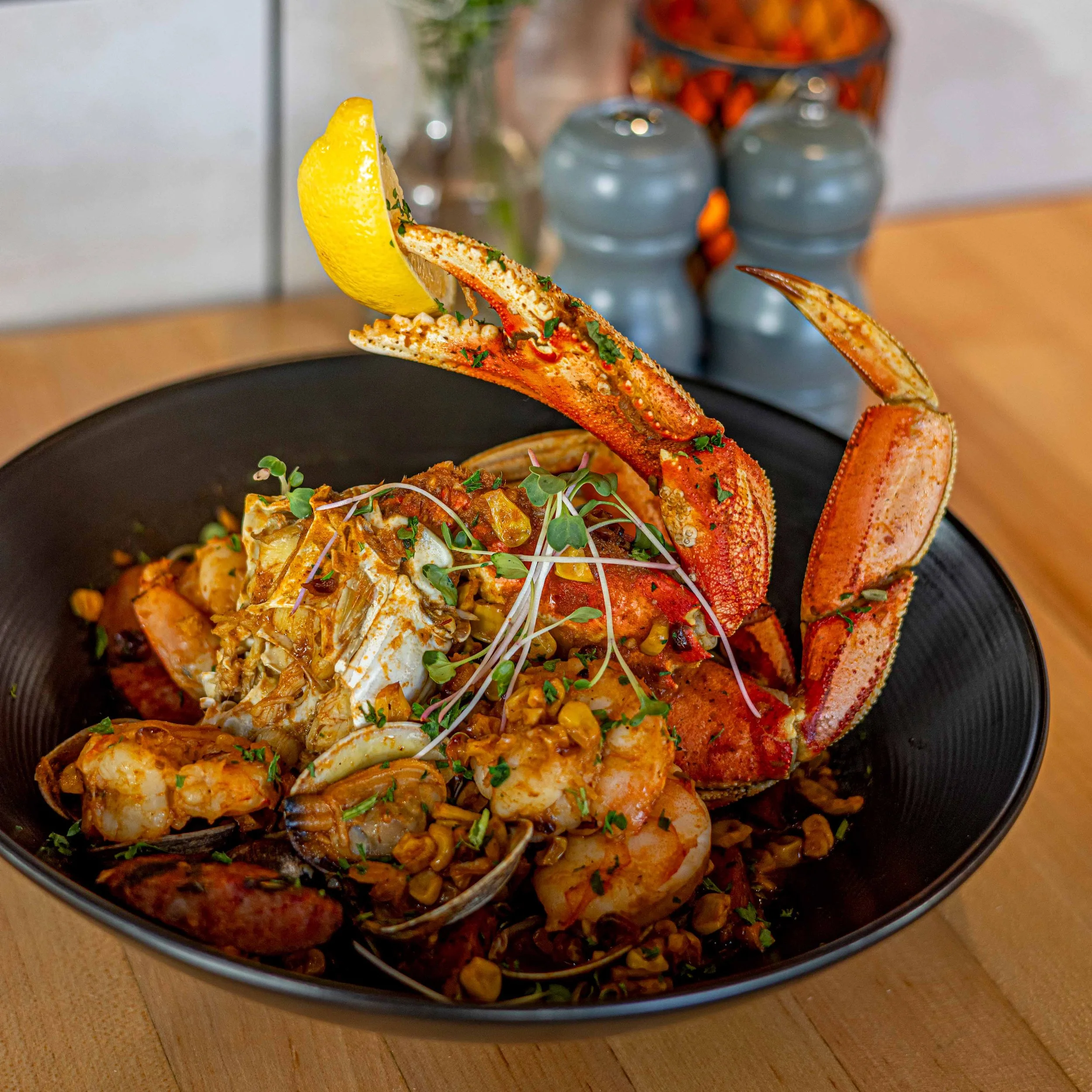 Dungenous crab dish with seafood, microgreens, and lemon wedge in a black bowl on a wooden table.