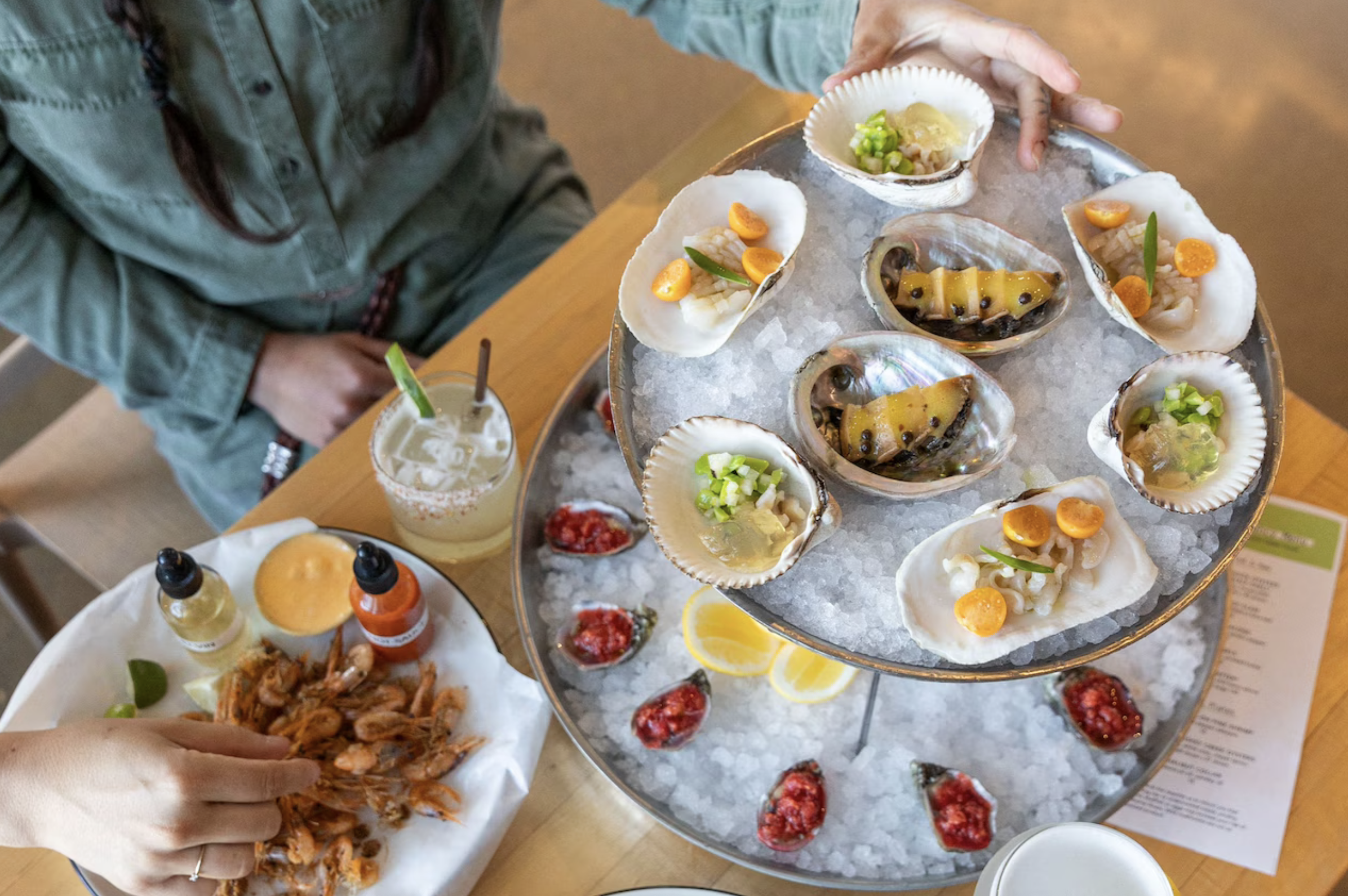 Seafood on a platter with oysters topped with chopped green onions, orange slices, and fresh herbs, set on a bed of ice. In the background, a person is reaching for seafood, and there are small bottles of hot sauce, a drink, and other dishes on the table.