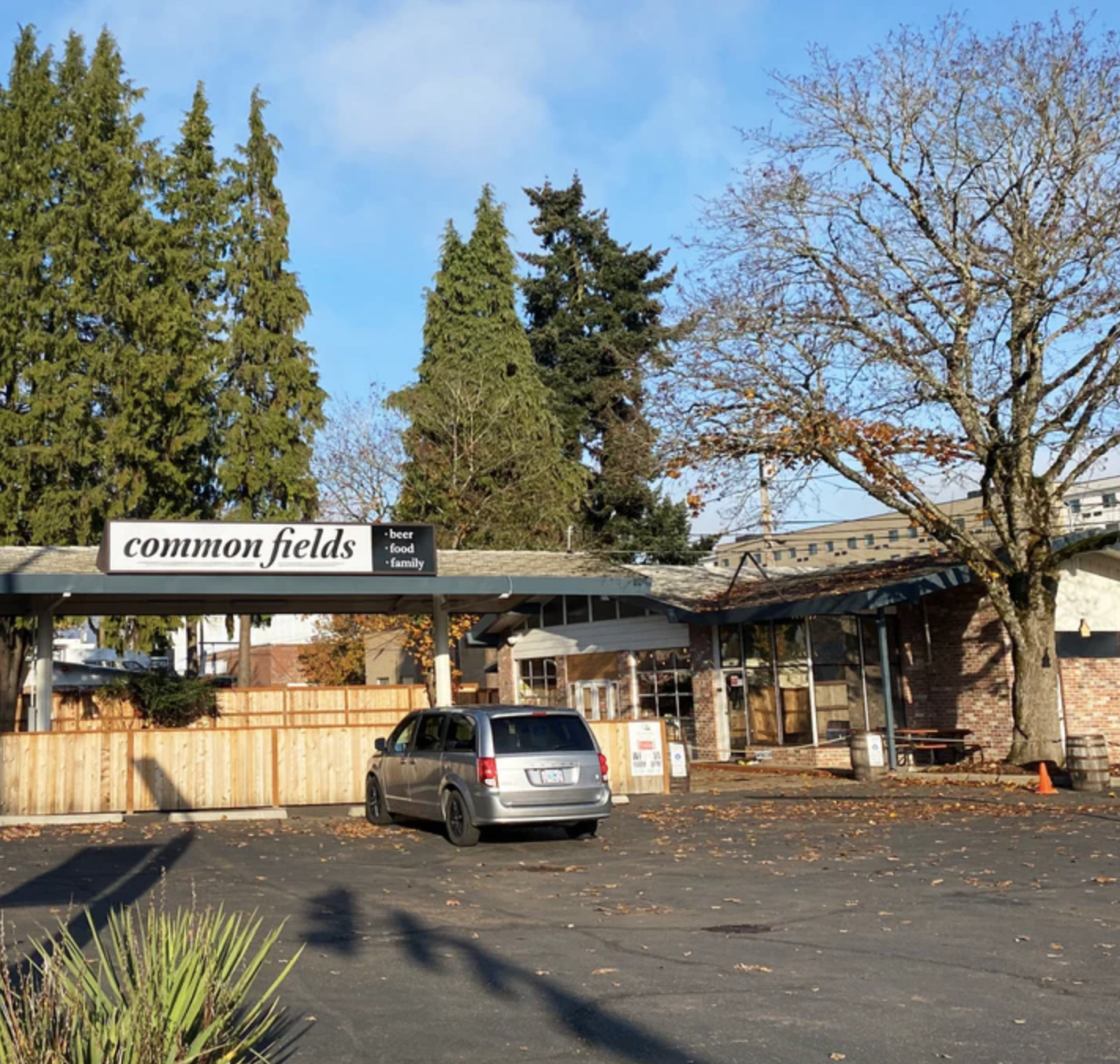 Exterior of Common Fields, a brick building with a parking area and a car, surrounded by trees and a wooden fence.