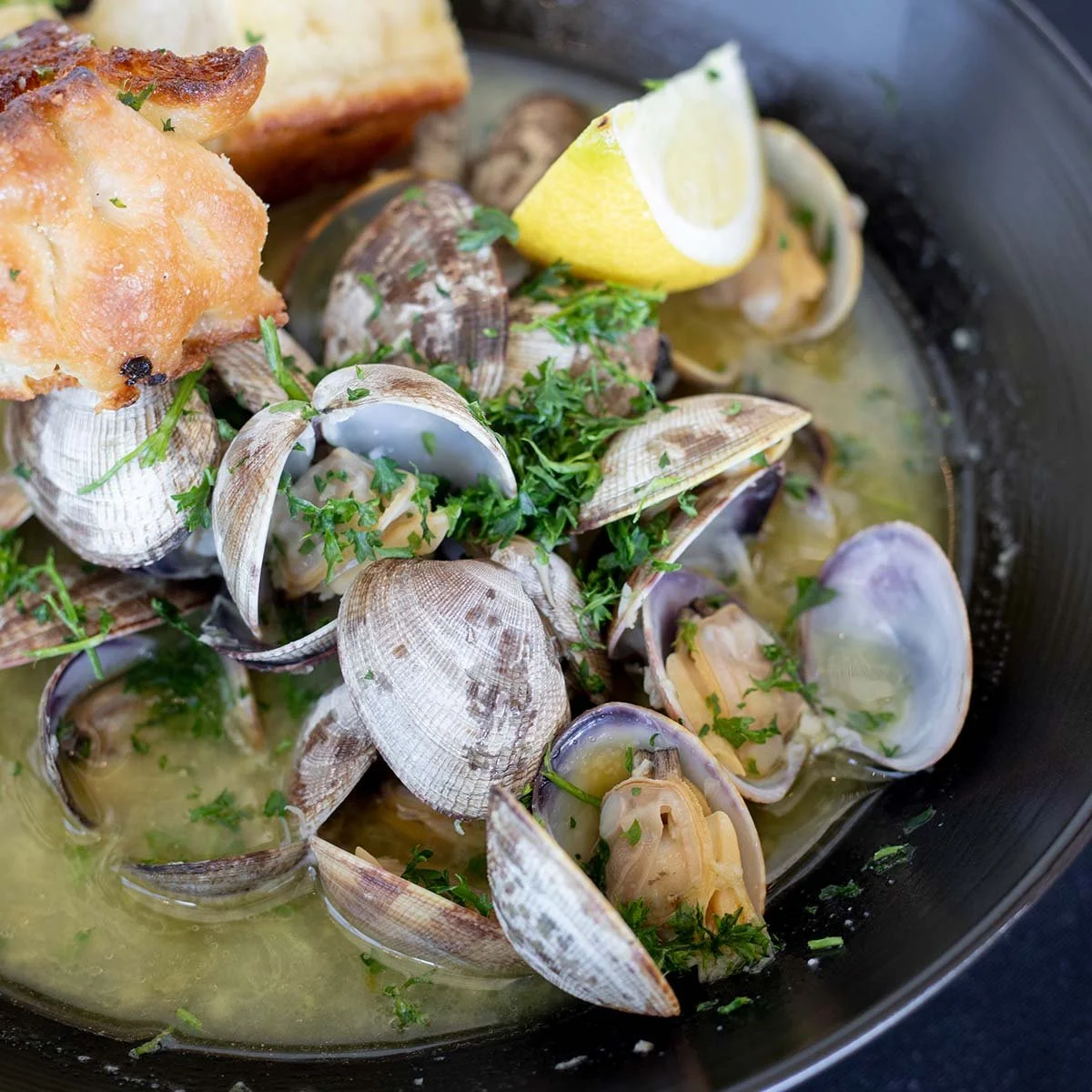 Clams in a broth garnished with chopped parsley and a lemon wedge.