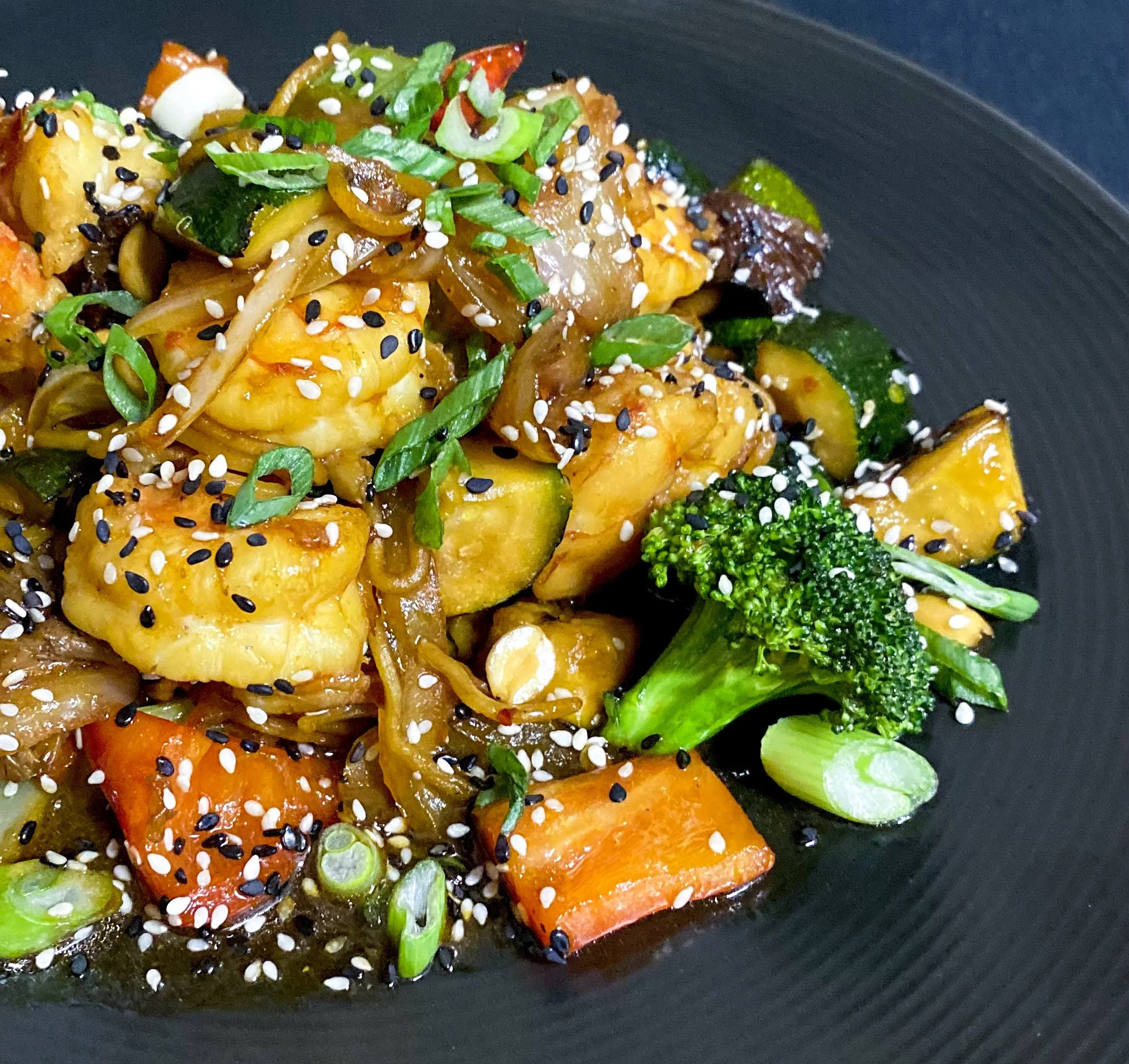 A black plate of stir-fried vegetables including broccoli, carrots, zucchini, and onions, topped with sesame seeds and chopped green onions.