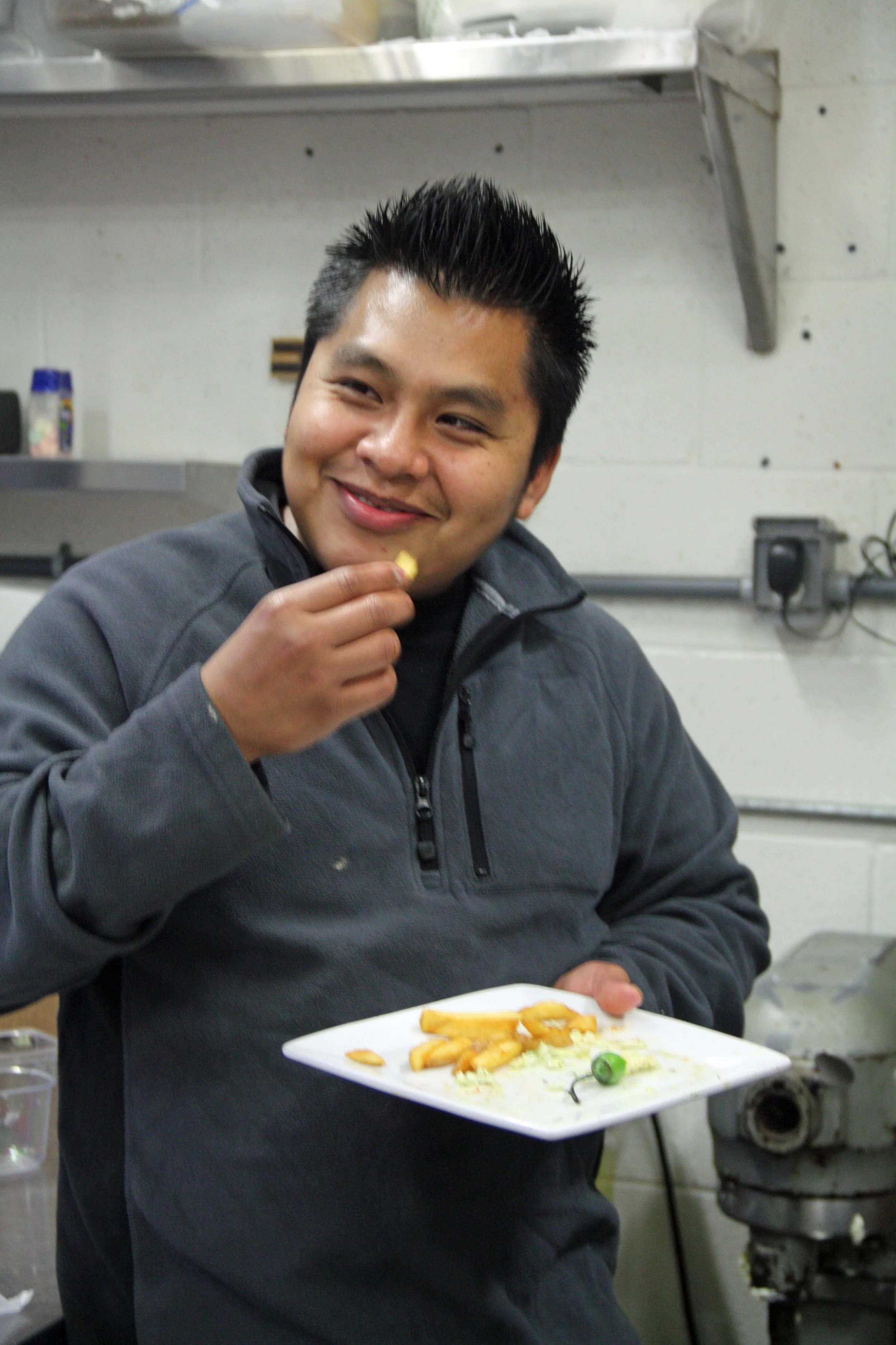 A young man with black spiked hair holding a white plate with some french fries, a green chili, and some lettuce, smiling in a kitchen setting.