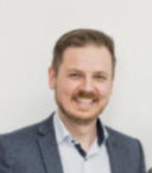 Ein lächelnder Johannes Katheder mit Bart in einem grauen Anzug vor neutralem Hintergrund.