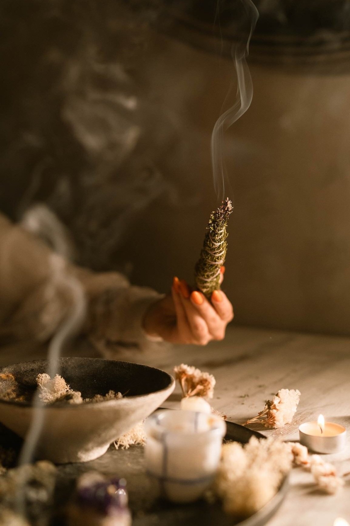 A person holding a smoldering sage bundle over a table with candles and dried flowers, with smoke rising from the sage.