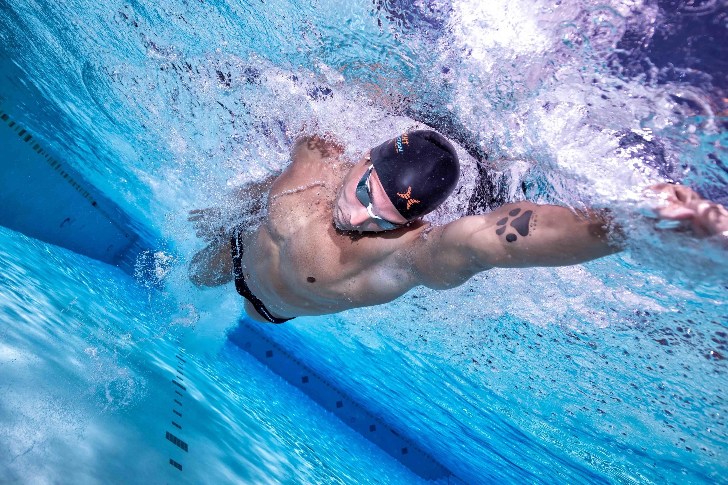Male swimmer in a black swim cap and goggles swimming freestyle in a pool, with a tattoo of paw print on his arm.