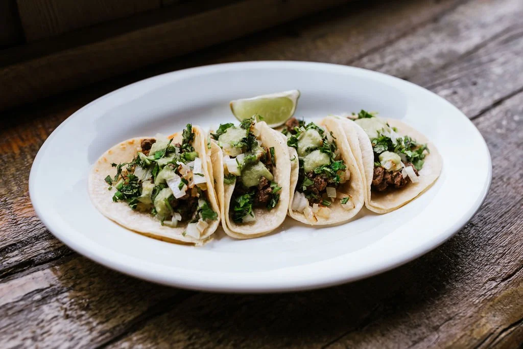 Four tacos filled with chopped beef, lettuce, and onions on a white plate with a lime wedge, on a wooden table.