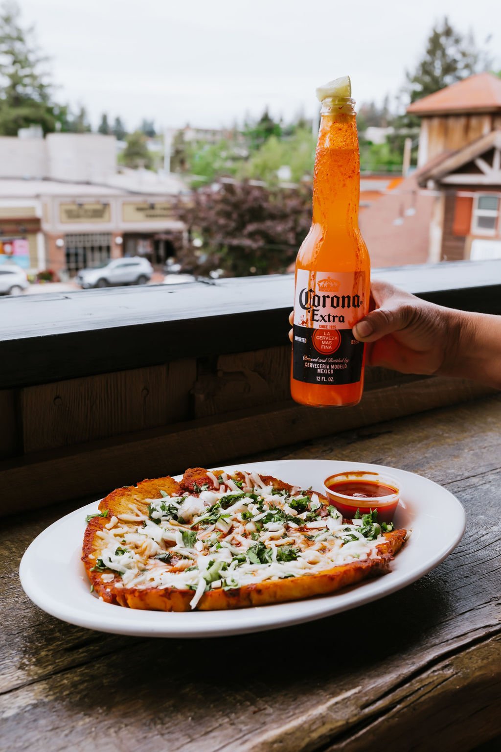 A hand holding a bottle of Corona Extra beer with a lime wedge, a pizza with cheese, vegetables, and sauce on a plate, and a small cup of red sauce on a wooden table indoors, with a view of a town street outside.