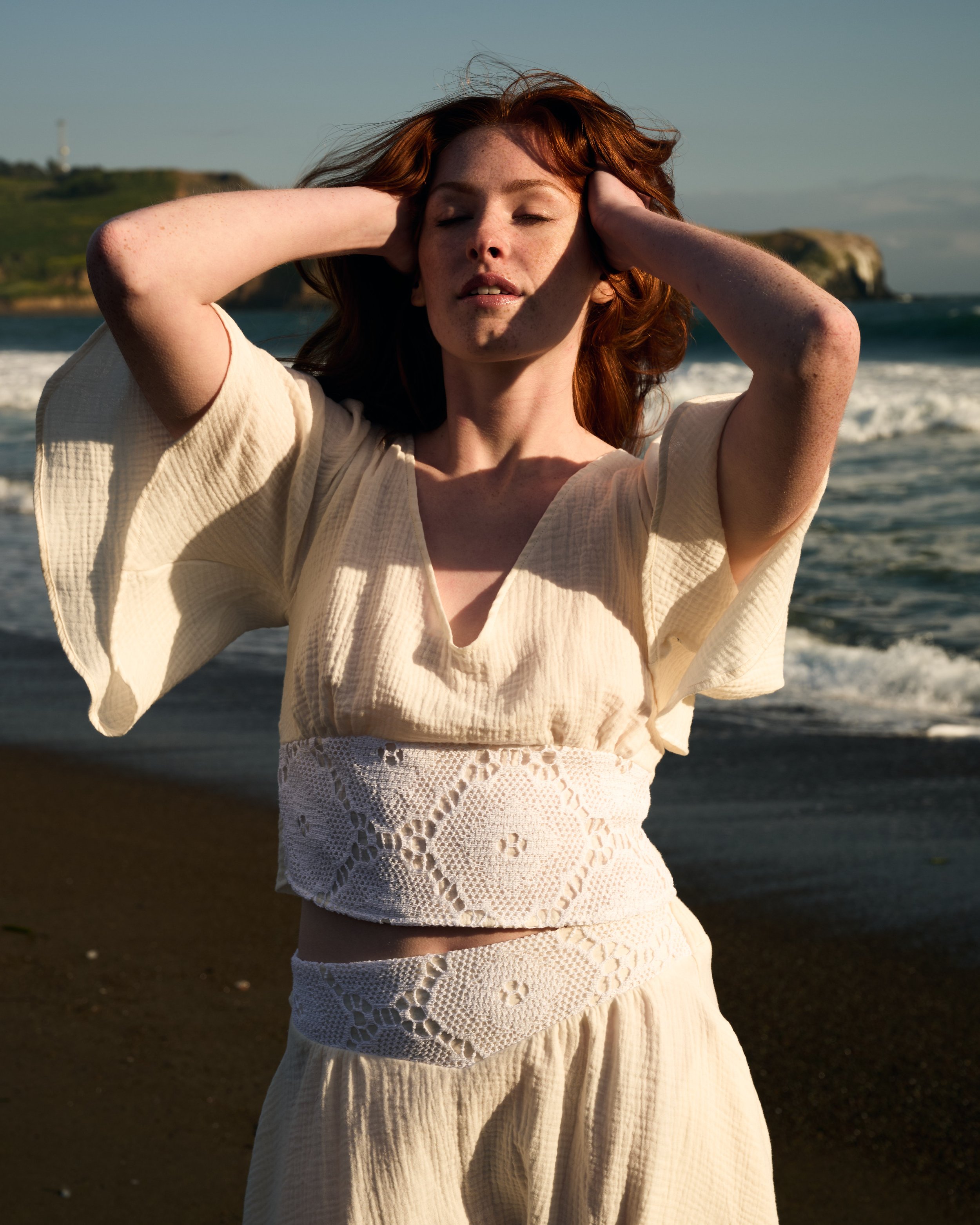Woman with red hair in a cream dress standing on a beach, eyes closed, hands on her head, sunny day.